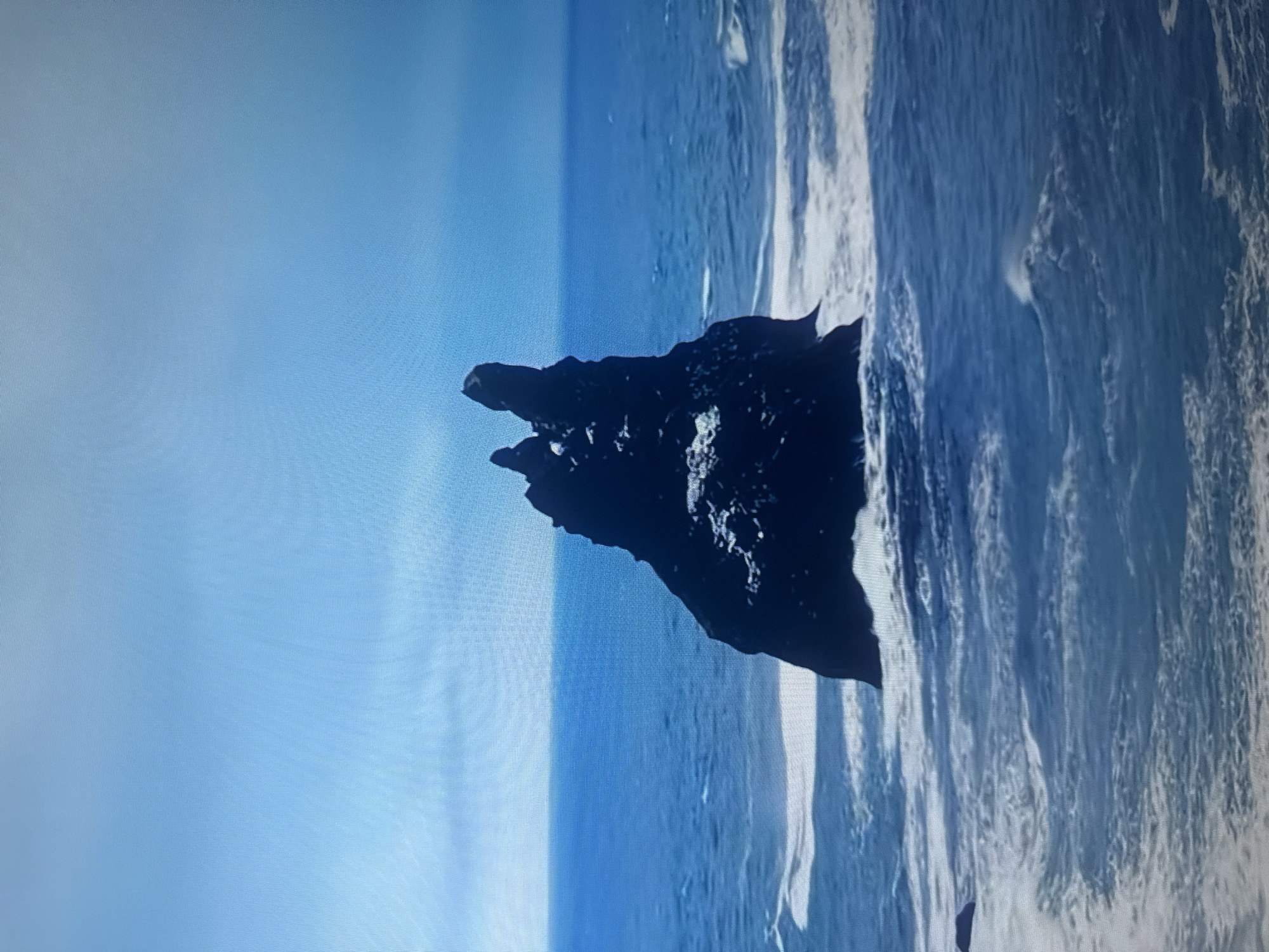 This is an image of a dark, jagged rock formation jutting out of the sea. The rock is covered in white, possibly snow or foam. The sea is a deep blue, with white waves crashing against the rock and breaking into foam. The sky is a clear, pale blue, with a slight gradient towards white at the top. The image appears to be taken from a distance, looking across the water towards the rock. There are no people or any signs of human activity. The location of this rock formation is not in Almaty, Kazakhstan, as Almaty is a landlocked city.
