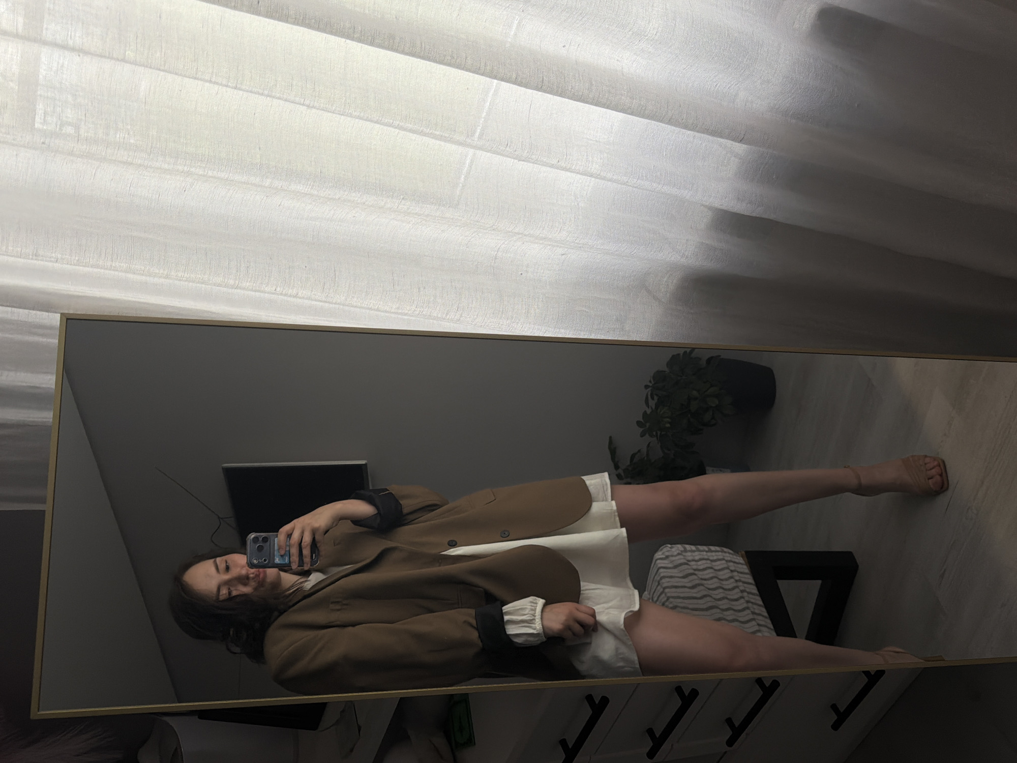 A woman is taking a mirror selfie in an indoor environment that appears to be a bedroom. She is wearing an oversized brown blazer over a white dress and beige strappy heels. The woman is holding a smartphone in her right hand to capture the reflection. Behind her, a dresser with black handles is visible, and to the right, sheer white curtains hang in front of a window. The room is dimly lit, with some natural light filtering through the curtains. There is a plant in a dark pot to the right of the dresser, and a framed mirror on the left side of the image. The overall setting suggests a domestic space. The specific location in the city of Chaikovsky, Russia, cannot be determined from the visual information provided in the image.