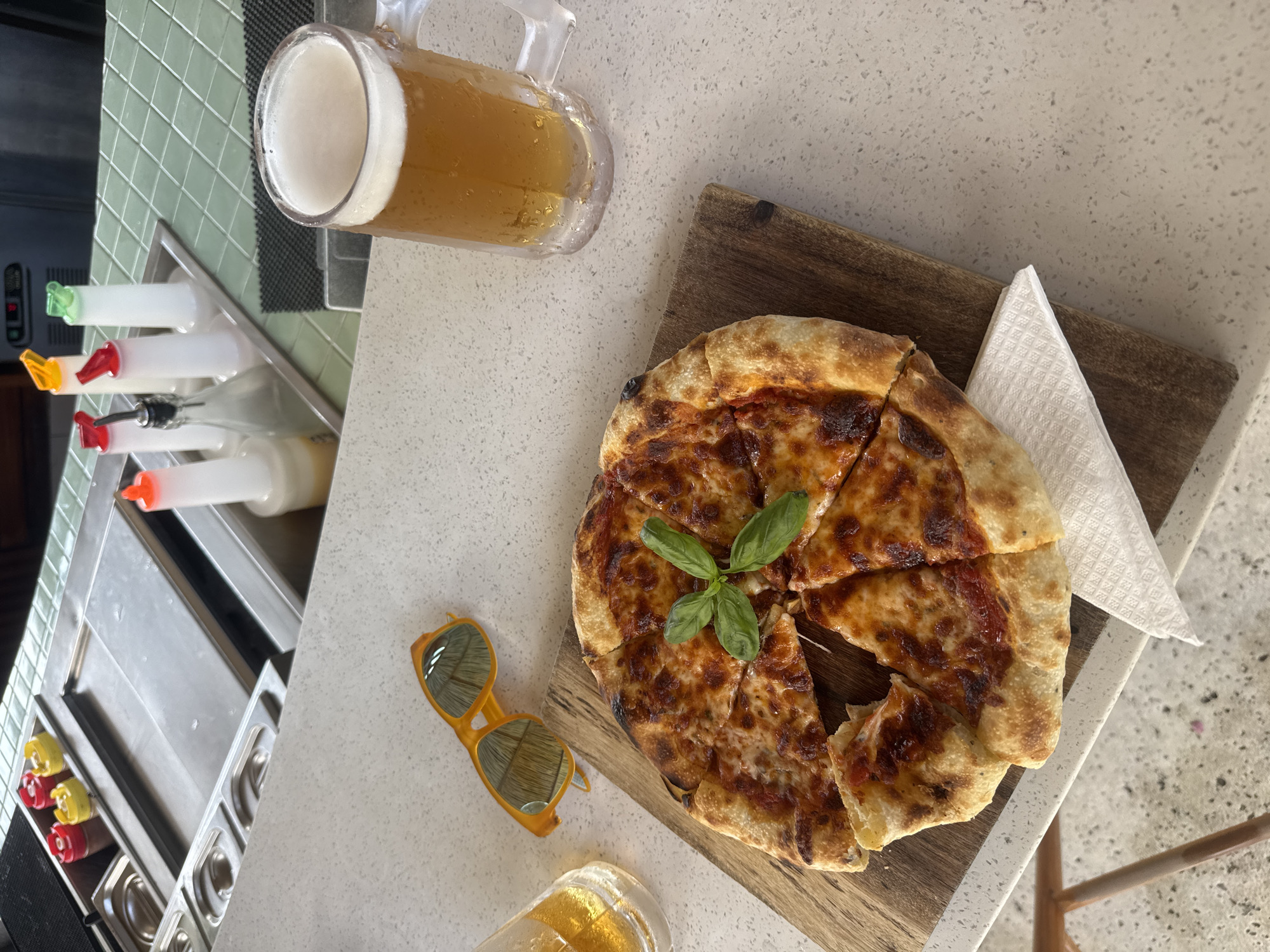 On a speckled, light-colored countertop, likely indoors, a high-angle shot displays a casual dining scene. The countertop could be made from granite or a similar material.
A partially consumed pizza, sliced into segments, sits on a rectangular wooden board. Fresh basil leaves garnish the center of the pizza. To the right of the pizza is a neatly folded white napkin.
To the upper left of the pizza, a clear glass beer mug is filled with a light-colored beer, its head of foam visible. Near the beer mug are a row of clear plastic bottles with colored caps.
Below the pizza and to the left are yellow-framed sunglasses, possibly reflecting the ambient light. A second, nearly empty beer mug rests near the sunglasses.
Based on the presence of beer and pizza, this is a casual dining scene. While not definitively identifiable, the setting resembles a restaurant or bar. Given the query about Kuta, Indonesia, the setting could reasonably be assumed to be a restaurant in that location.