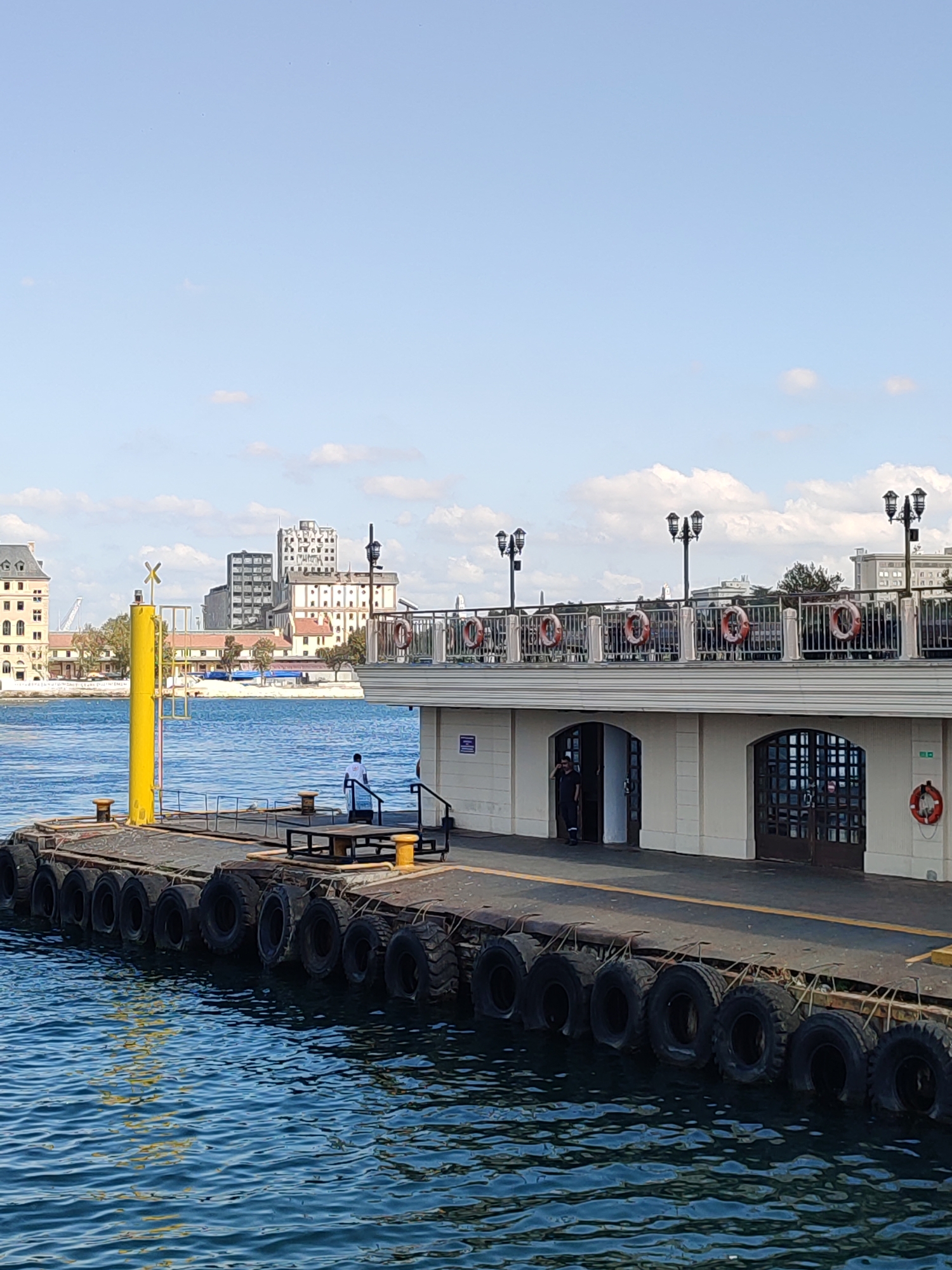 This image depicts a pier in Kadıköy, Turkey, under a bright, partly cloudy sky. The pier extends from the foreground into the mid-ground, with the calm, blue water of the Sea of Marmara surrounding it. The pier itself is lined with numerous large tires, serving as fenders, some partially submerged in the water.
In the foreground, a yellow post with an "X" marking is visible, along with mooring posts. Further along the pier, there is a seating area and a person in a white shirt standing. In the mid-ground, a two-story structure is situated on the pier. The lower level features archways, one of which has two individuals standing near the entrance, and the other appears to be a closed doorway with glass panels. The upper level of this structure is a walkway with a railing decorated with several life preservers. Three ornate lampposts are spaced along the walkway.
In the background, across the water, the cityscape of Kadıköy is visible. This includes several buildings of varying architectural styles and heights, some appearing to be older, industrial structures with multiple windows, and others more modern. The overall scene suggests a functional port area, likely used for ferry services or other maritime activities, on a clear day.