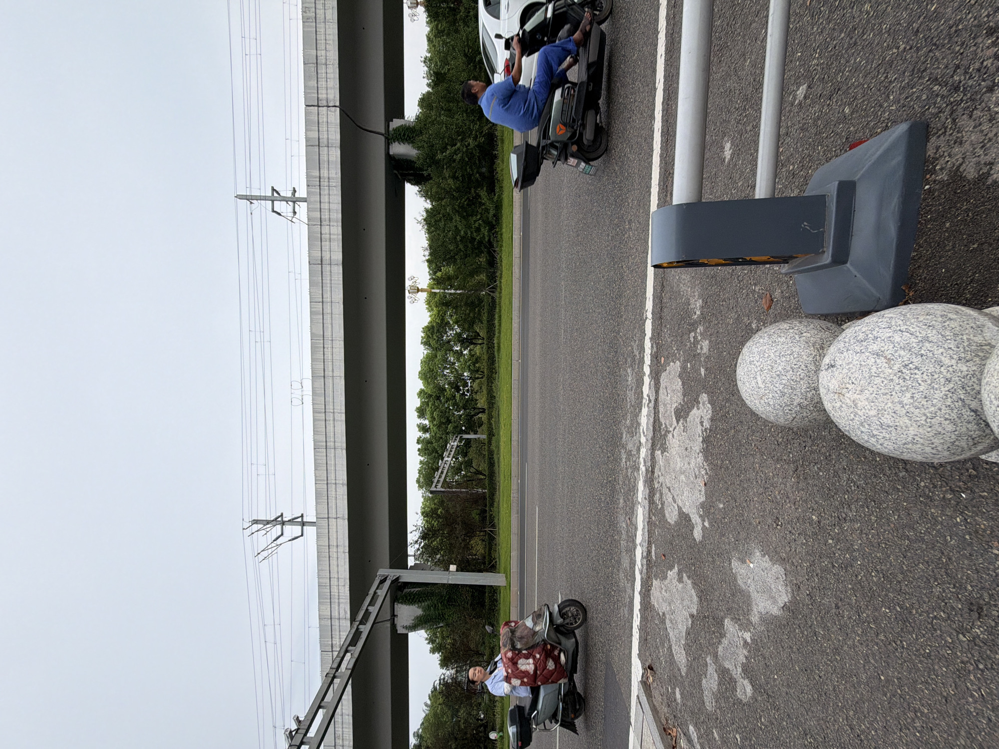 An outdoor, eye-level, medium shot captures a street scene in Jiangjin, China, under an overcast sky. Two individuals on electric scooters navigate the asphalt road, which is bordered by a grassy verge and dense green trees. Above the trees, a concrete overpass supports overhead power lines and what appears to be a railway track. In the foreground, two large, speckled spherical bollards stand beside a traffic barrier. The scooter on the left is ridden by a person wearing a light blue shirt and a patterned blanket. The scooter on the right is ridden by a person in a blue jumpsuit. A white car is partially visible behind the scooter on the right. The road surface exhibits some dark, damp patches.