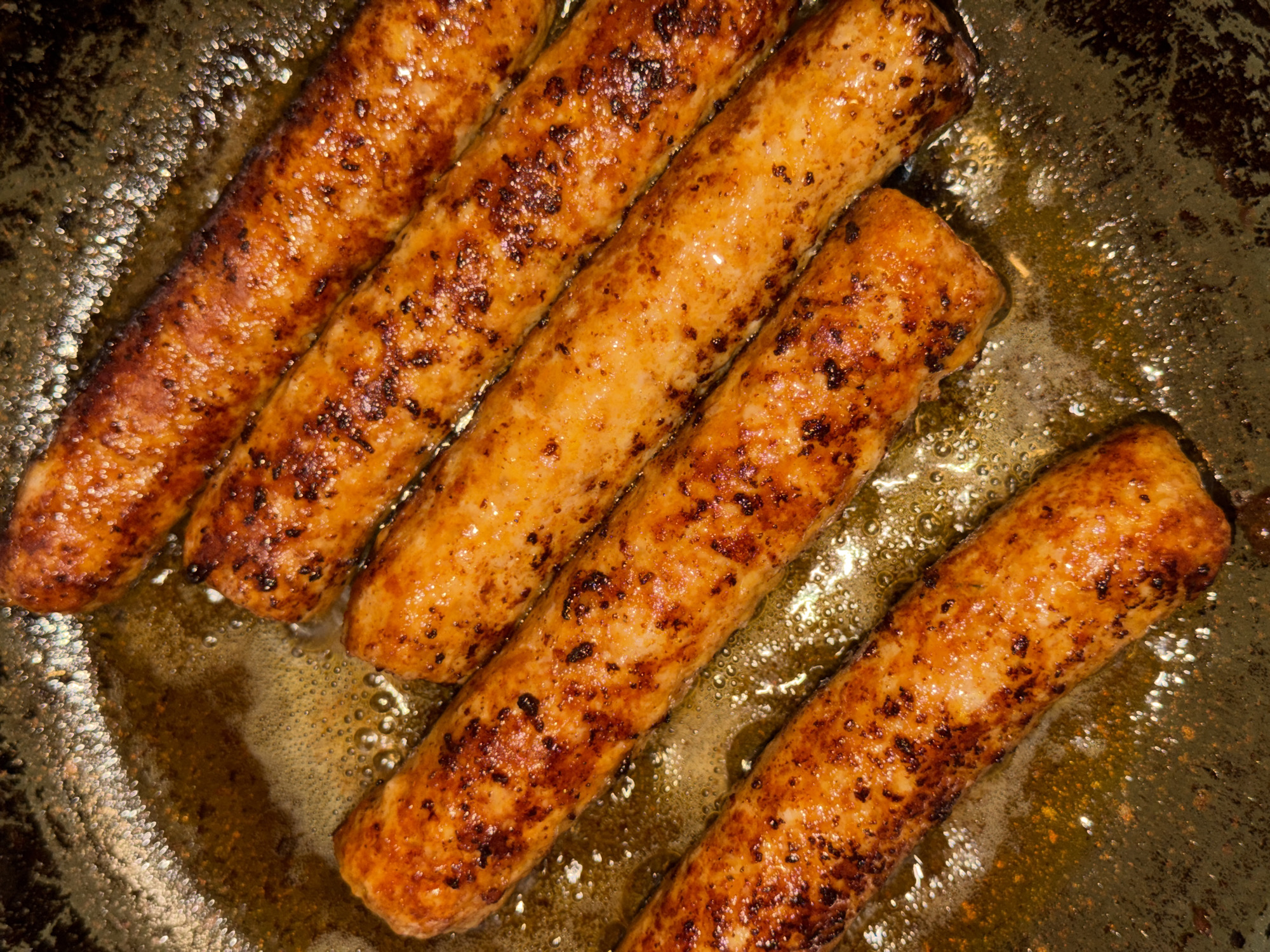 This is a close-up, overhead shot of six sausages cooking in a pan. The sausages are golden brown with darker, charred bits, indicating they are well-seared. They are arranged diagonally in the pan, with the top sausage positioned near the top right corner of the frame. The pan contains a significant amount of glistening oil, with visible bubbles forming around the sausages. The pan itself appears to be dark and possibly seasoned, with some texture visible along the edges. The lighting creates highlights on the oil and the surface of the sausages. There are no people visible in the image. The scene is a kitchen or food preparation area. There is no information within the image to indicate the location is in the city of Khimki, Russia.
