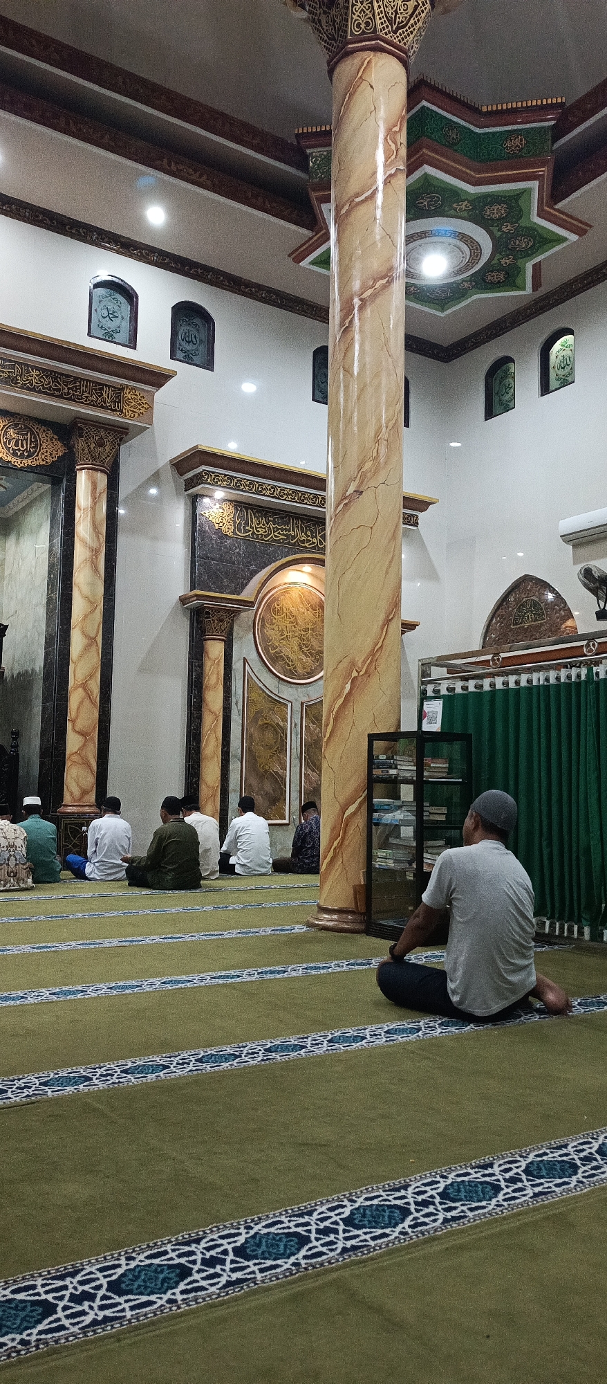 The image depicts the interior of a mosque, specifically the Arjasa Mosque in Arjasa, Indonesia. Several men are seated on a green carpet with decorative blue and white striped borders, facing a wall adorned with Islamic calligraphy and decorative elements. A large, marble-patterned pillar is prominent in the foreground, supporting a ornately decorated ceiling. A shelf unit filled with books stands near the pillar. The overall atmosphere suggests a place of worship during a prayer or gathering.