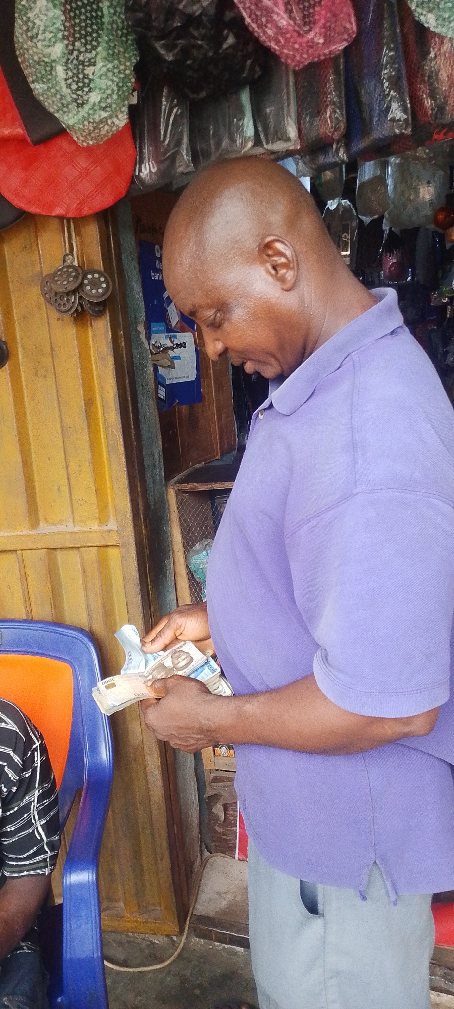 A middle-aged man with a shaved head wearing a lavender short-sleeved polo shirt and light-colored pants is counting a stack of Nigerian Naira notes. He is standing in what appears to be a small shop or kiosk. The shop has a yellow-painted door with visible vertical panels and several items hanging from the doorframe and above, including what appears to be some kind of metal tools and various packaged goods. A blue sign with text that includes "bank" is visible on the door. A blue chair with an orange back is partially visible on the left side of the image, with a person wearing a striped shirt seated on it. The location is likely in a marketplace or busy commercial area of Lokoja, Nigeria, based on the nature of the shop.