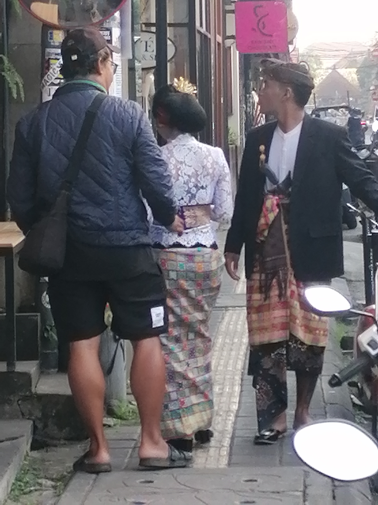In Ubud District, Indonesia, three individuals are walking on a paved sidewalk next to a building. The person on the left, wearing a blue quilted jacket, dark shorts, and a baseball cap, is walking away from the camera. To their right, a person in a white lace top and a patterned sarong walks forward, their back to the camera. A third person, dressed in a black blazer over a white shirt and a colorful sarong, walks ahead of them, looking to their right. A motorcycle is parked on the right side of the image. The environment suggests an urban or semi-urban street scene with foliage visible in the background.
