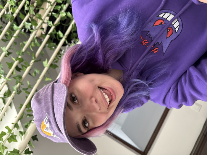 An indoor, close-up selfie shows a young adult female from the chest up, smiling widely and extending her tongue. She has multi-toned hair, primarily purple with pink highlights, and wears a light purple baseball cap featuring a yellow crescent moon and white mountain peaks embroidered on the front. Her attire includes a purple hooded sweatshirt with a prominent embroidered image of the Pokémon character Gengar and Japanese text on the chest. The background consists of a light-colored wall with a visible light switch or outlet to the left, a dark brown door frame behind her, and a light-colored trellis decorated with artificial green ivy leaves to the right. The specific location within the City of Zagreb, Croatia, cannot be determined from the provided image.