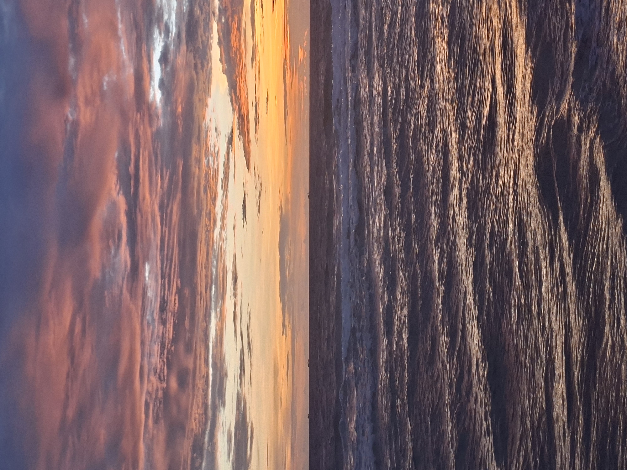 A sunset over the ocean creates a dramatic sky with hues of pink, orange, and purple. The clouds are textured and layered, reflecting the colors of the setting sun. Below the sky, the ocean is a deep, muted purple, with gentle waves creating ripples and highlights. A few distant boats are visible on the horizon, silhouetted against the bright sky. The scene is serene and captures the beauty of a coastal sunset. This image is likely taken from a beach or a coastal viewpoint in Phu Quoc, Vietnam, a popular destination known for its stunning sunsets and ocean views.