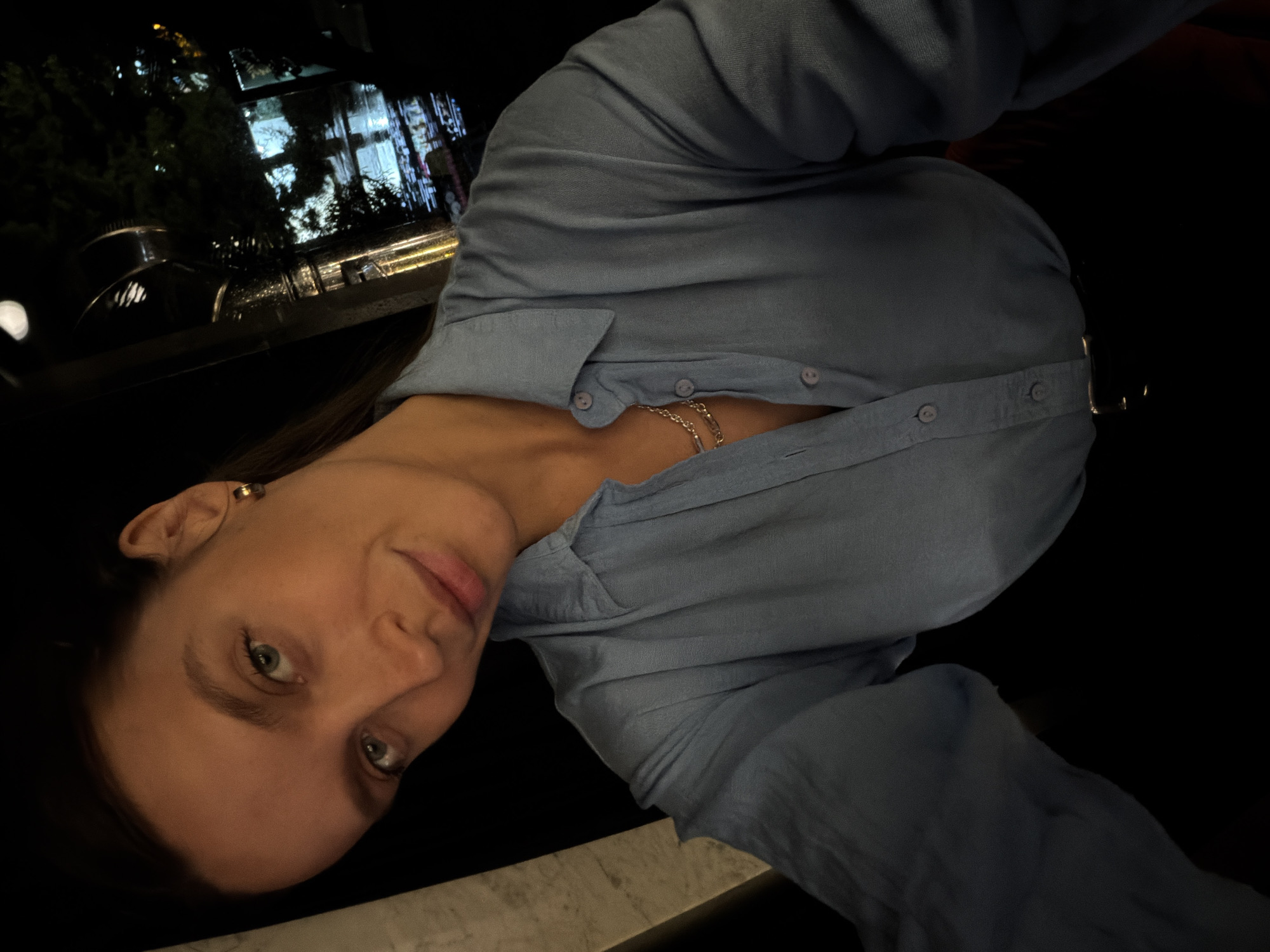 A low-light, tilted portrait-oriented image captures a young woman at night, likely in an outdoor setting or a space with large windows overlooking a city scene. She gazes directly at the camera with a calm and slightly engaging expression, her light-colored eyes and medium-brown hair framed by the dim lighting. She is wearing a light blue or denim-colored collared shirt, unbuttoned to reveal a gold-toned layered necklace. A small gold ear cuff adorns her right ear.
The background is dark, indicative of nighttime, with soft, out-of-focus reflections of artificial lights from distant buildings, streetlights, and possibly trees, suggesting an urban or semi-urban environment. These reflections appear on a dark, reflective surface, hinting at a window, glass railing, or even a body of water. The woman appears to be leaning against a light-colored, possibly stone or concrete, railing or ledge, which forms the bottom edge of the frame. The overall atmosphere is intimate, softly lit by ambient sources, consistent with an evening in a location like Budva, Montenegro. No visible text is present.