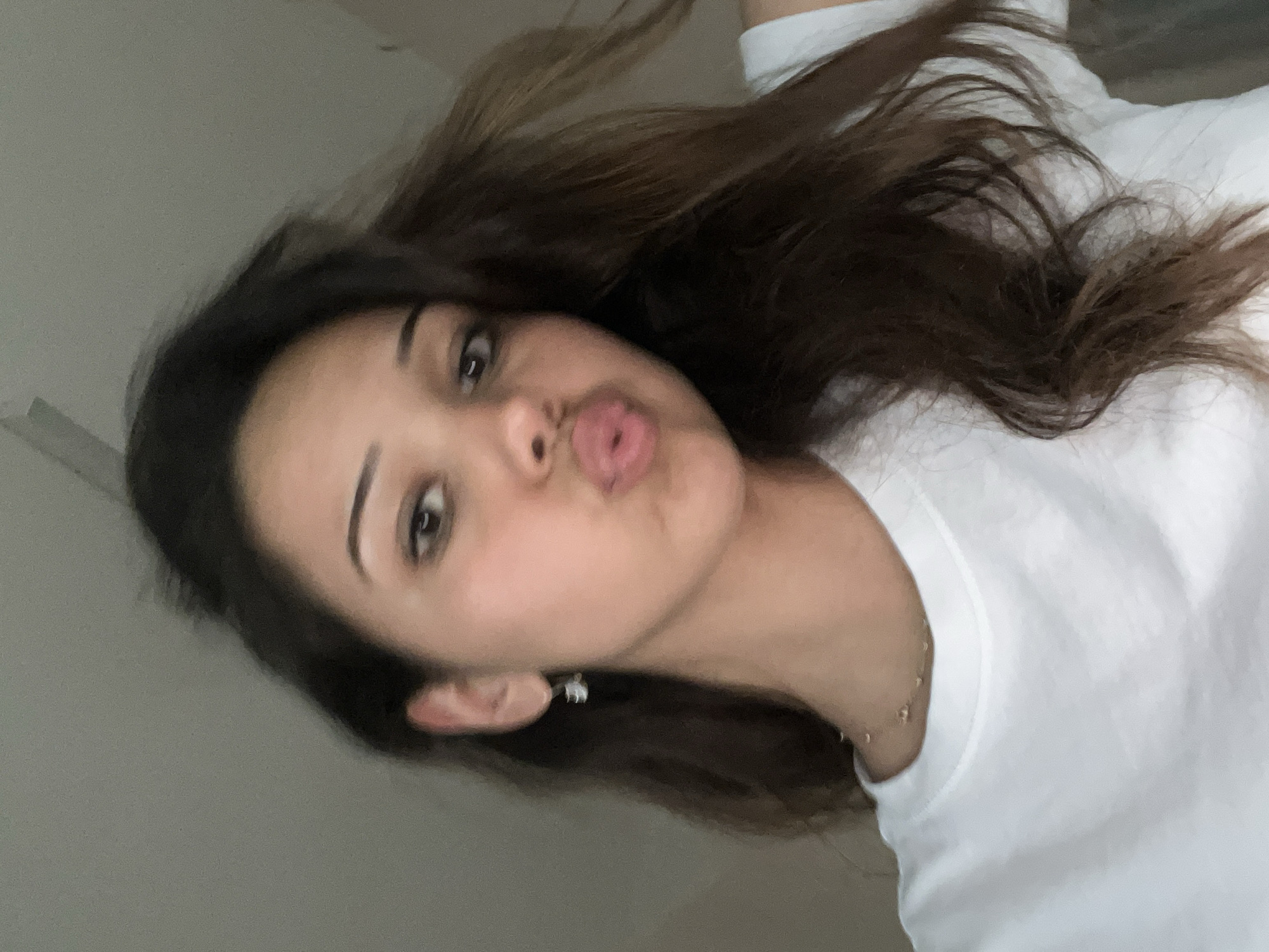 The image is a close-up selfie of a young woman with dark hair and brown eyes. She is wearing a white t-shirt and a delicate gold necklace. Her lips are pursed in a kiss, and she is wearing a small stud earring in her left ear. The background is a plain, light-colored wall, suggesting an indoor setting. There are no discernible elements in the image that would place the scene in Zlatograd, Bulgaria.