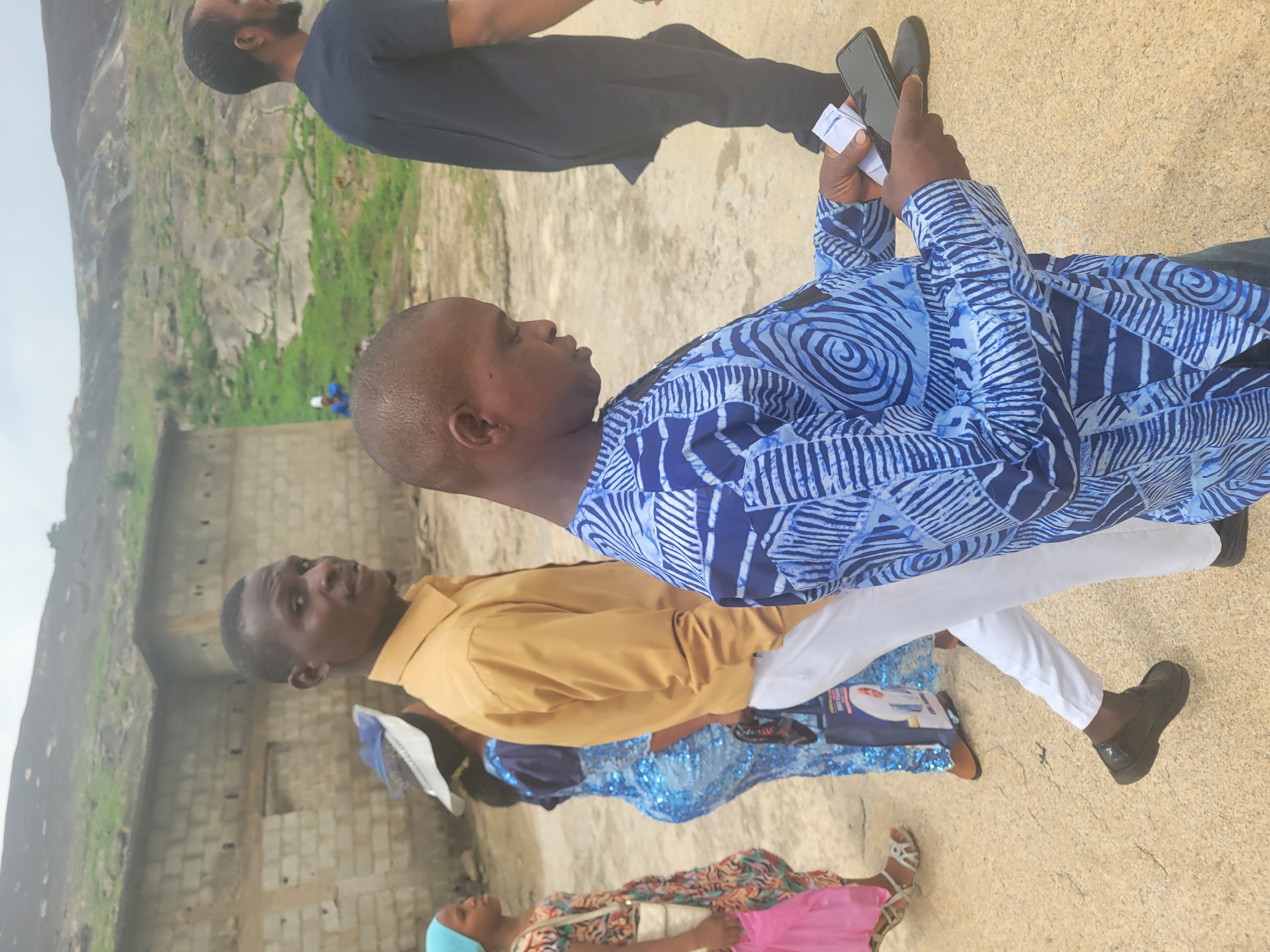 The image depicts a group of people outdoors. One man is in the foreground, wearing a blue patterned shirt and white trousers, holding a cell phone and what appears to be a couple of small white tickets or cards. To his left and slightly behind him is another man in a yellow shirt. Further back, there is a woman wearing a colorful patterned dress with a pink element and a head covering.
The setting appears to be near a stone or concrete structure, possibly a wall, and in the background, there is a large rock formation. Based on the location provided, the image seems to be taken in Ibo, Nigeria. People are standing, there is no visible interaction beyond their presence in the same space.