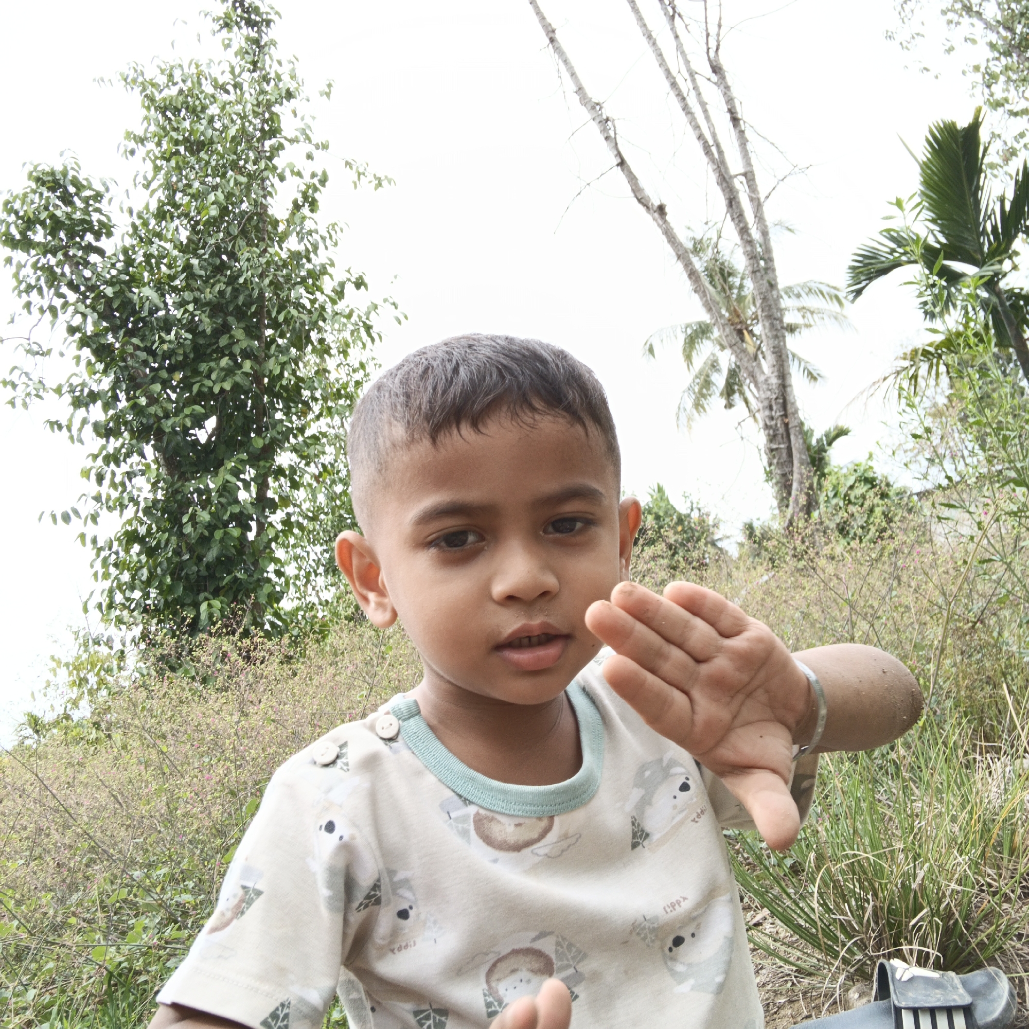 This is a close-up, medium shot portrait of a young boy, likely of Indonesian descent, outdoors in what appears to be a rural setting in Geunteng, Indonesia. The boy is facing the camera, his gaze directed forward with a neutral expression. His hair is dark and short. He is wearing a light-colored t-shirt with a repeating pattern of cartoon koala bears and geometric shapes. His right arm is raised, hand open, with fingers spread, and he is wearing a thin, silver bracelet on his wrist. The background is lush with vegetation, featuring various trees and plants, some with small pinkish flowers. The sky is overcast and bright. The overall lighting suggests it is daytime.