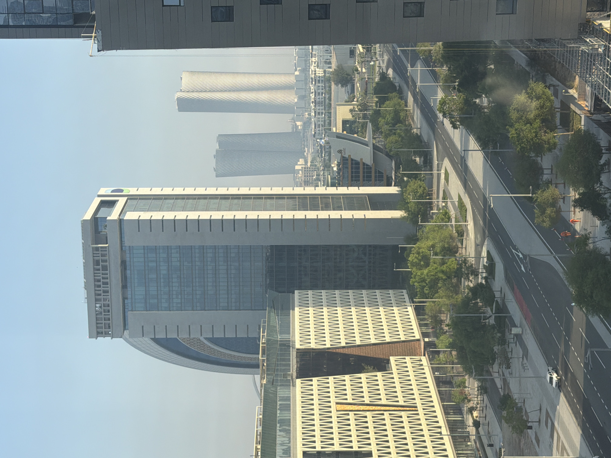 An aerial view captures a modern city scene under a clear sky. In the foreground, a white SUV drives on a multi-lane road lined with trees and streetlights. Adjacent to the road are two large buildings: a beige one with a distinctive patterned facade and a tall, gray one. Behind these, a tall, light-colored building with a curved top and a glass facade is visible. Further in the distance, two strikingly twisted, white skyscrapers stand out against the hazy horizon, alongside other urban structures and low-rise buildings. The scene is bathed in daylight, suggesting it is daytime.
