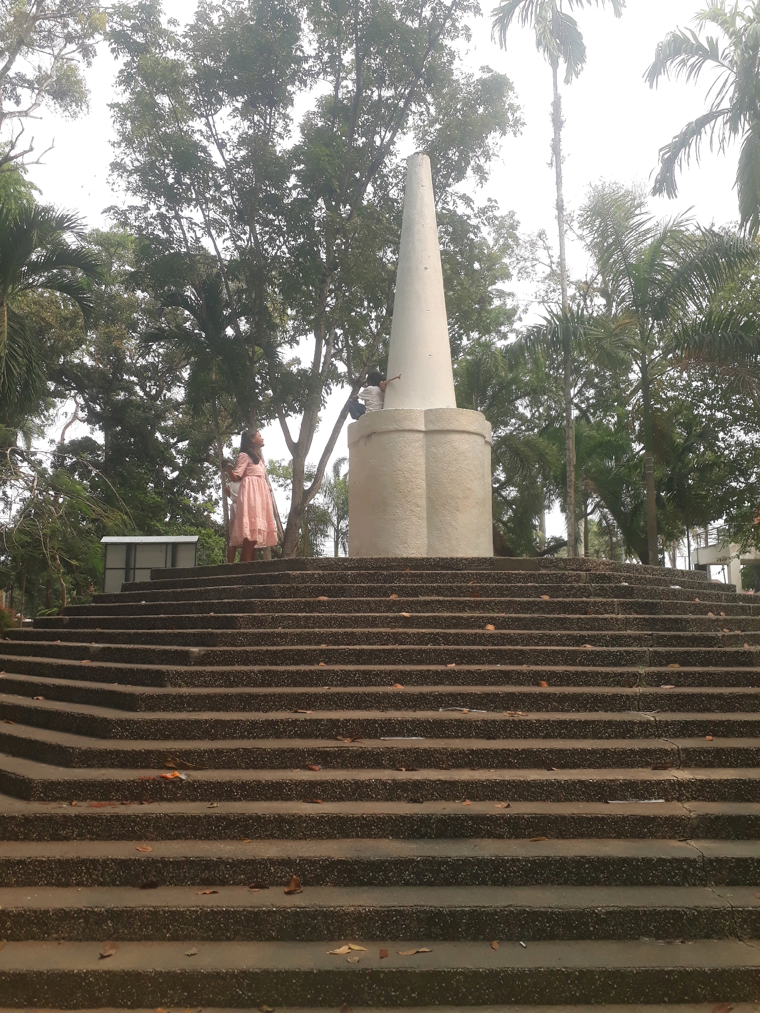 A young girl wearing a pink dress stands on a wide, tiered staircase leading up to a large, conical monument. The monument is light-colored and sits atop a broader, octagonal base. Behind the monument, lush green trees and palm trees create a dense backdrop. The sky is overcast. In the background, to the left of the monument and slightly behind the girl, a small, gray, shed-like structure is visible. Another person, wearing a white shirt, is partially visible on the right side of the monument's base, pointing upwards. The staircase is made of stone with a speckled texture and is littered with fallen leaves. The scene is likely located in a park or public space in Pangkalpinang, Indonesia, given the tropical vegetation and monument.