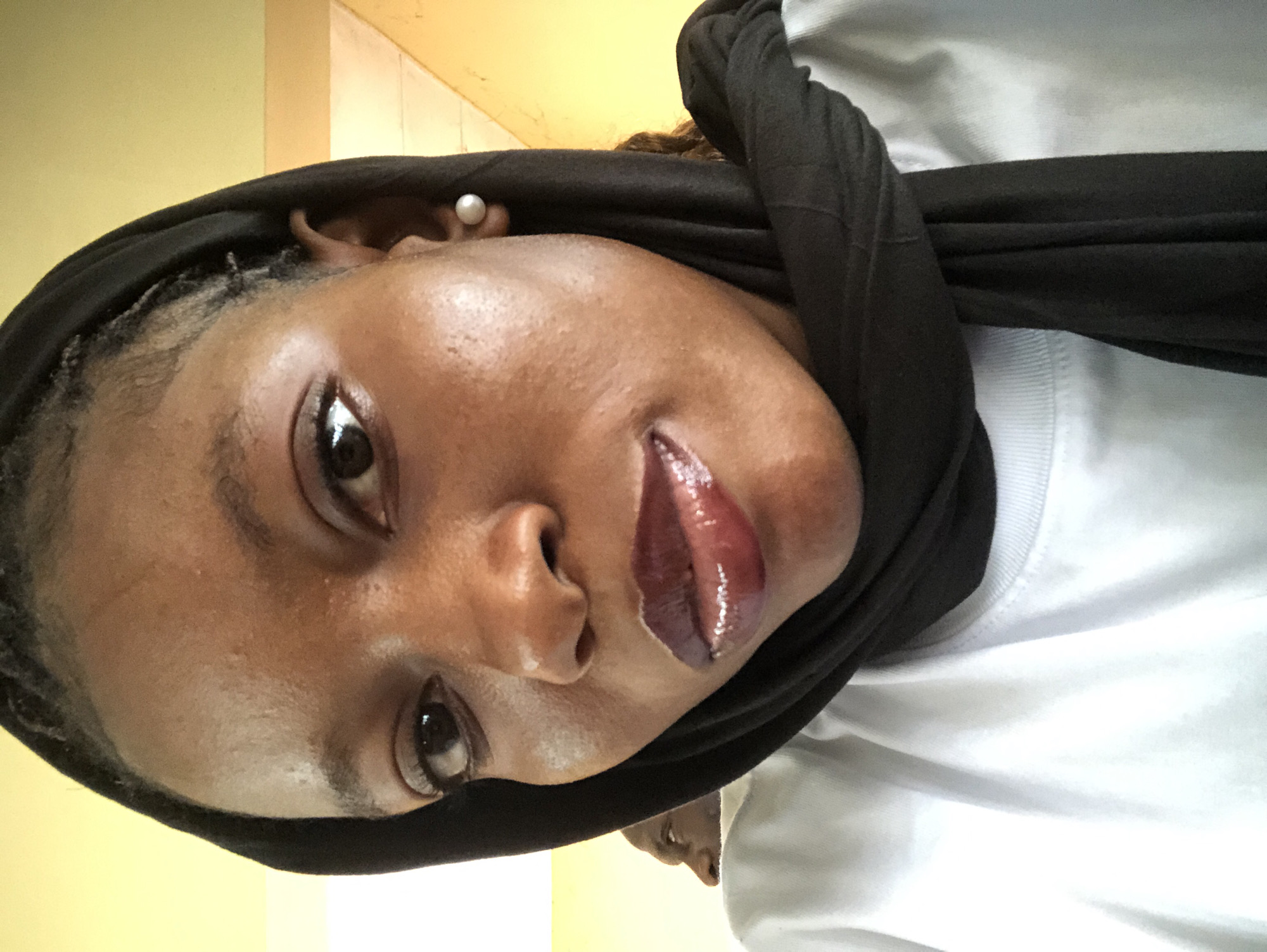A close-up photograph shows a dark-skinned individual wearing a black headscarf and a white t-shirt. The person has dark eyes and glossy, dark-colored lips. An earring is visible on the right ear. The background consists of a pale yellow wall. There is no verifiable information to indicate this image was taken in Pindiga, Nigeria.