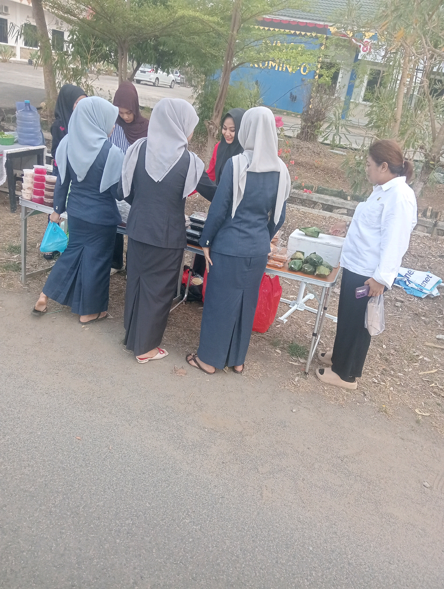 The image shows a group of people, mostly women, standing around two folding tables set up outdoors. The setting appears to be a street or sidewalk, with a building in the background and trees and dry vegetation surrounding the area. The building in the background has blue walls with yellow and white signage. One of the tables is laden with containers of food items, possibly for sale, including what looks like drinks in large plastic bottles, and small red and clear containers with colorful contents. The other table has a cooler box with several food items wrapped in green leaves.
There are at least five women visible. Three of them are wearing dark blue or gray formal-looking outfits with headscarves. One woman in the foreground is wearing a white long-sleeved shirt and black pants, and she is holding a phone. Another woman, visible from behind, is wearing a dark blue headscarf. Two women are interacting with the food on the tables, and the woman in the white shirt appears to be observing or waiting. Red plastic bags and some shopping bags are also visible near the tables. The ground is paved and appears dusty, with some dry leaves and twigs scattered around. The overall impression is of an outdoor vendor or a small informal market. The presence of the building with signage suggests it could be a public area or a service building within Sidoharjo, Indonesia.