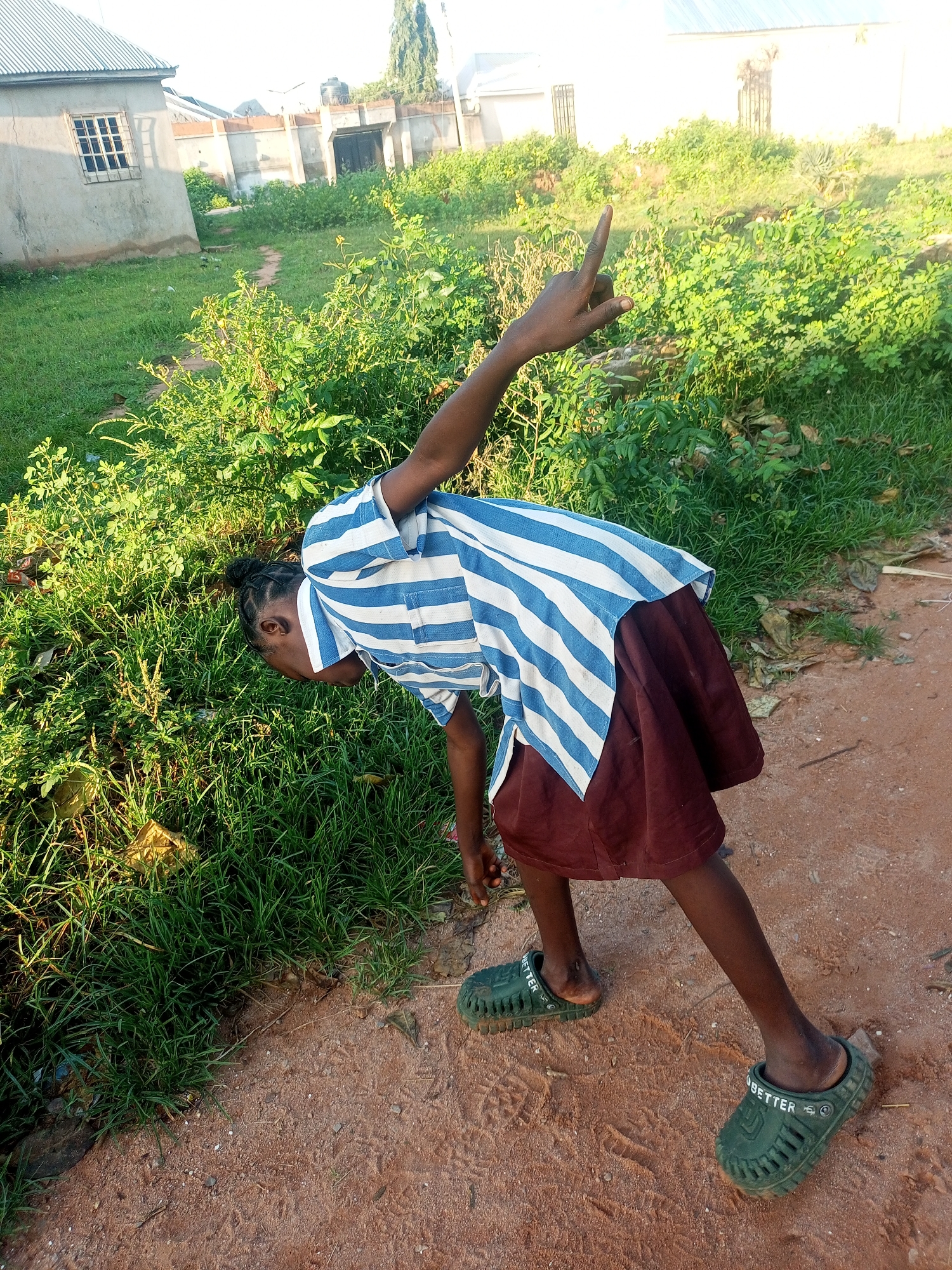 A young girl, dressed in a blue and white striped shirt and a maroon skirt, leans forward in a grassy outdoor area. She is wearing green slip-on shoes. Her right hand is raised with her index finger pointing upwards. Behind her, a dirt path leads towards a building with a metal grate window. Further in the background, there are more buildings and vegetation. The scene appears to be in a semi-rural or residential area within Tempure, Nigeria.