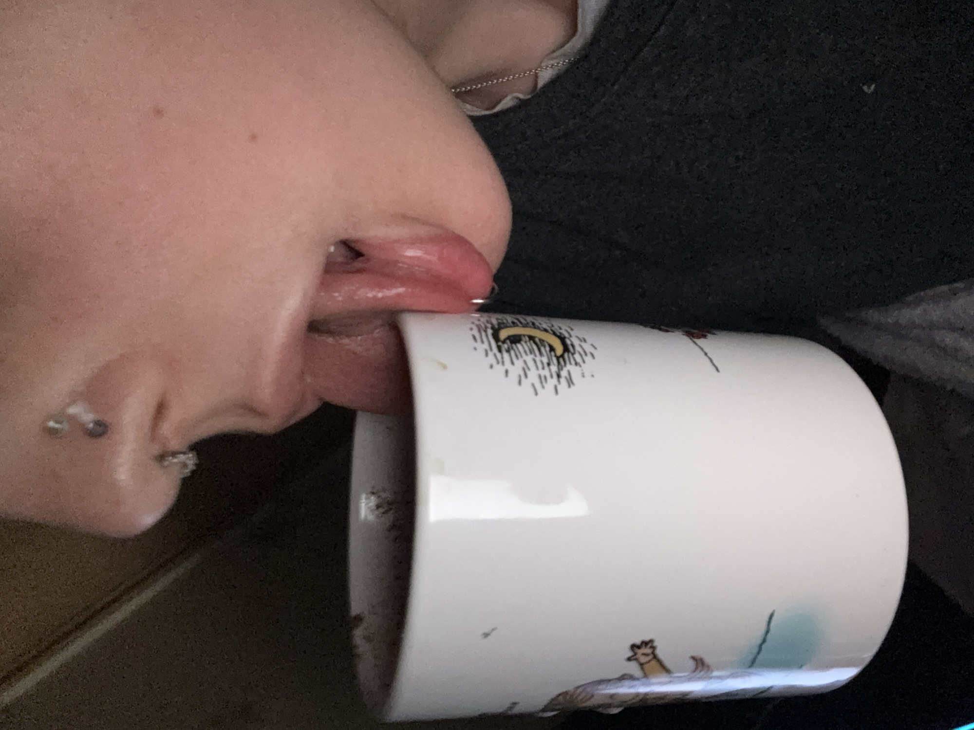 A close-up shot shows a person's face, with their tongue extended and seemingly licking the rim of a white mug. The person has multiple piercings on their ear and a piercing on their nose. They are wearing a gray top with a silver chain necklace visible. The mug has a cartoon-style illustration on it. No landmarks or details suggest a location in Moscow, Russia.