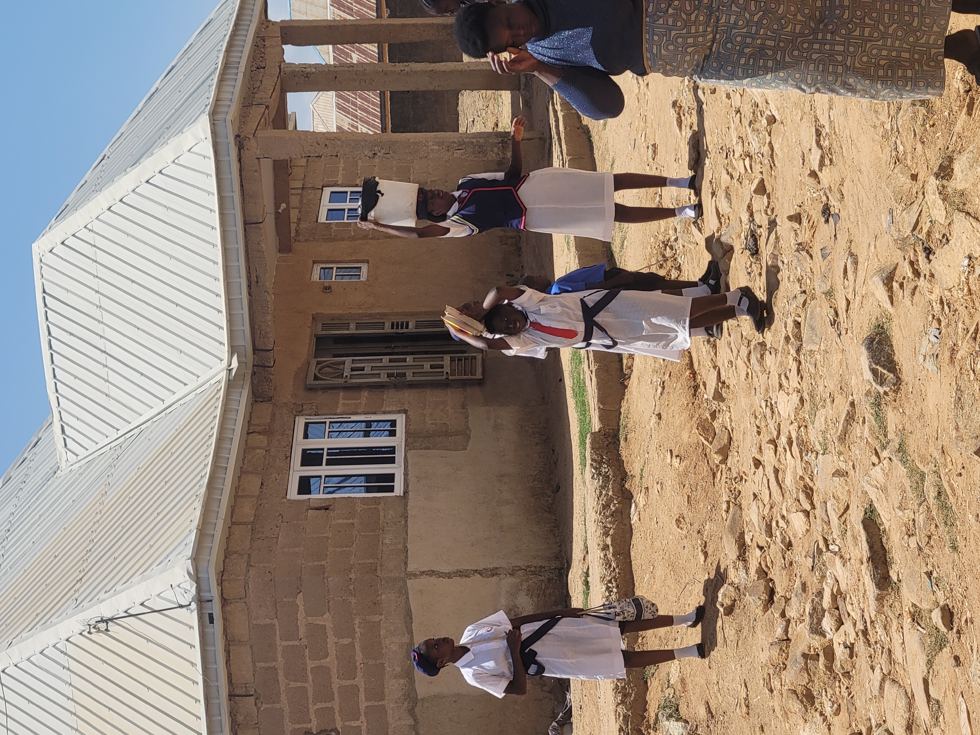 The image depicts a group of individuals in what appears to be a residential area. Three young individuals in school uniforms stand in front of a building constructed of brick or cinder blocks. They are holding what appear to be books or papers above their heads. The ground is dry and dusty. An adult individual stands in the background, partially visible. The building has a corrugated metal roof. The image was taken during daylight under clear skies. The location is consistent with the city of Jos, Nigeria.