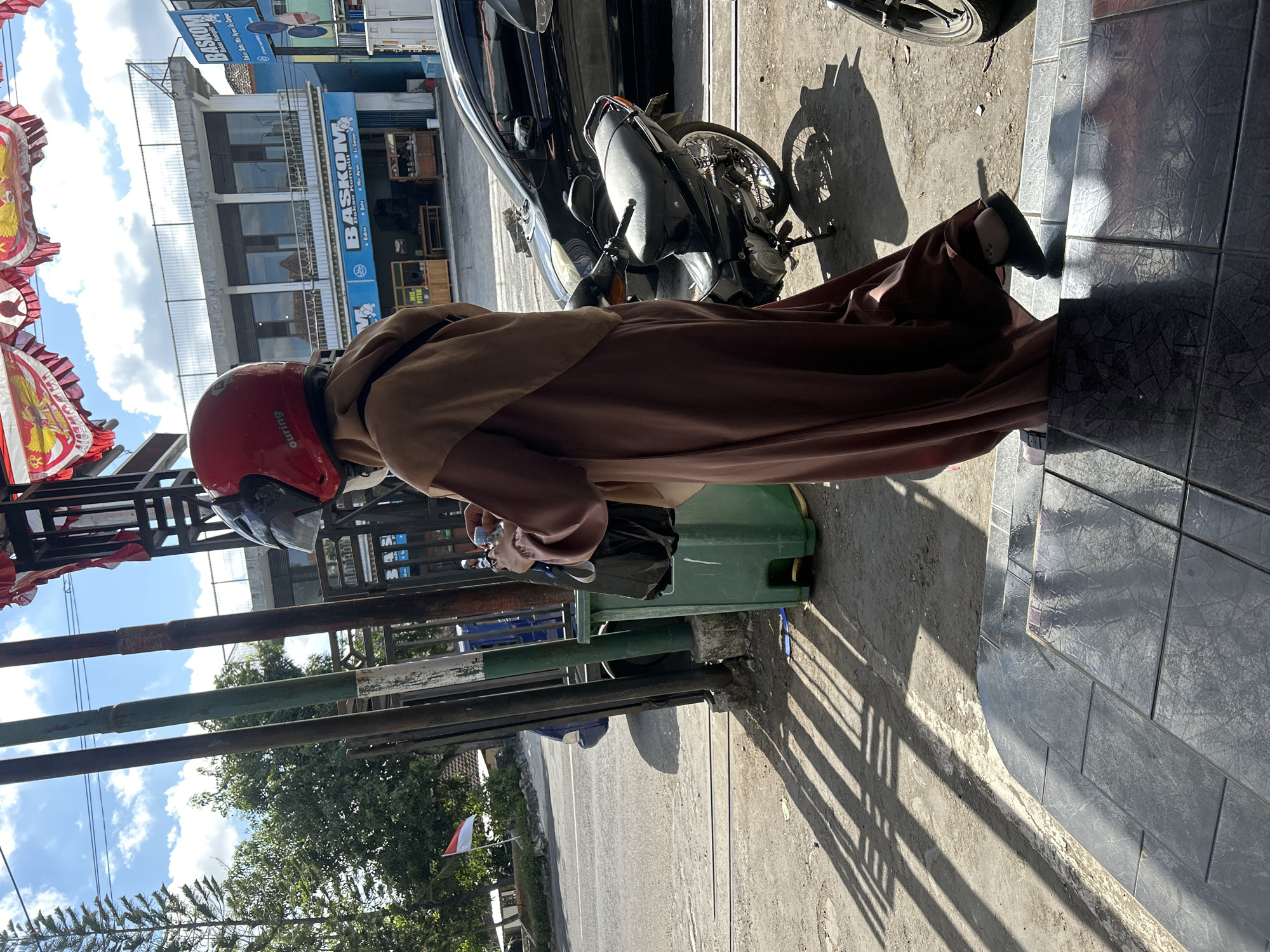 A person wearing a brown abaya, a light brown headscarf, and a red helmet walks away from the camera on a tiled walkway. The person is carrying a plastic bag in their right hand and a bottle of water in their left hand. In the background, there is a black motorcycle parked next to a black car. Behind the vehicles, a blue building with signage that reads "BASKOM" and "BASKOM KUSUS" is visible. A green trash can stands to the left of the person. The scene appears to be outdoors, possibly in a commercial area, with shadows from a fence cast across the ground. The sky is blue with scattered clouds. The Indonesian flag is visible on a pole in the distance. The location is identified as the city of Wonosari, Indonesia.
