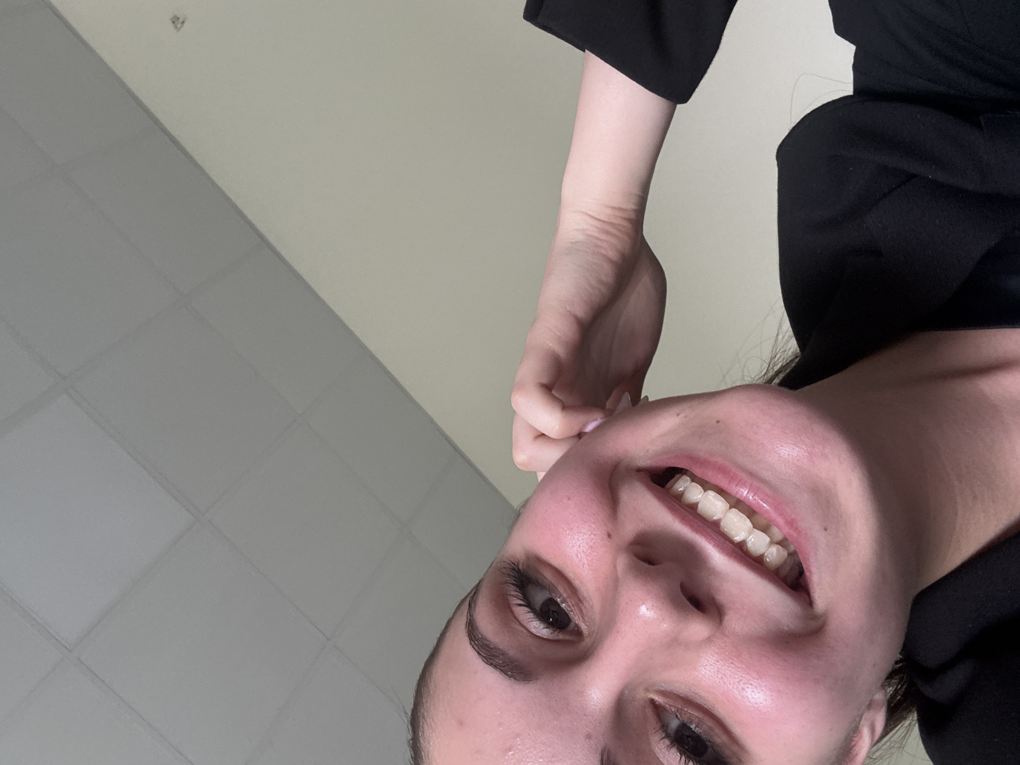 A low-angle, close-up selfie captures a young woman smiling broadly and looking down towards the camera. Her head is tilted, and her happy expression is evident through her wide smile, which reveals both upper and lower teeth fitted with clear dental aligners or a retainer. She has natural makeup, bright eyes, and well-defined eyebrows. Her light skin is visible, and she appears to be wearing a dark, possibly black, top or jacket. Her right arm is bent, with her hand positioned near her ear or neck.
The background consists of an indoor ceiling, diagonally divided. The upper right portion features a light, plain white ceiling, while the lower left portion shows a grid pattern of light grey square ceiling tiles. A small, indistinct dark object or mark is visible on the plain white ceiling section. The lighting suggests an interior setting. There is no visible text in the image. The location context is Burgas, Bulgaria.