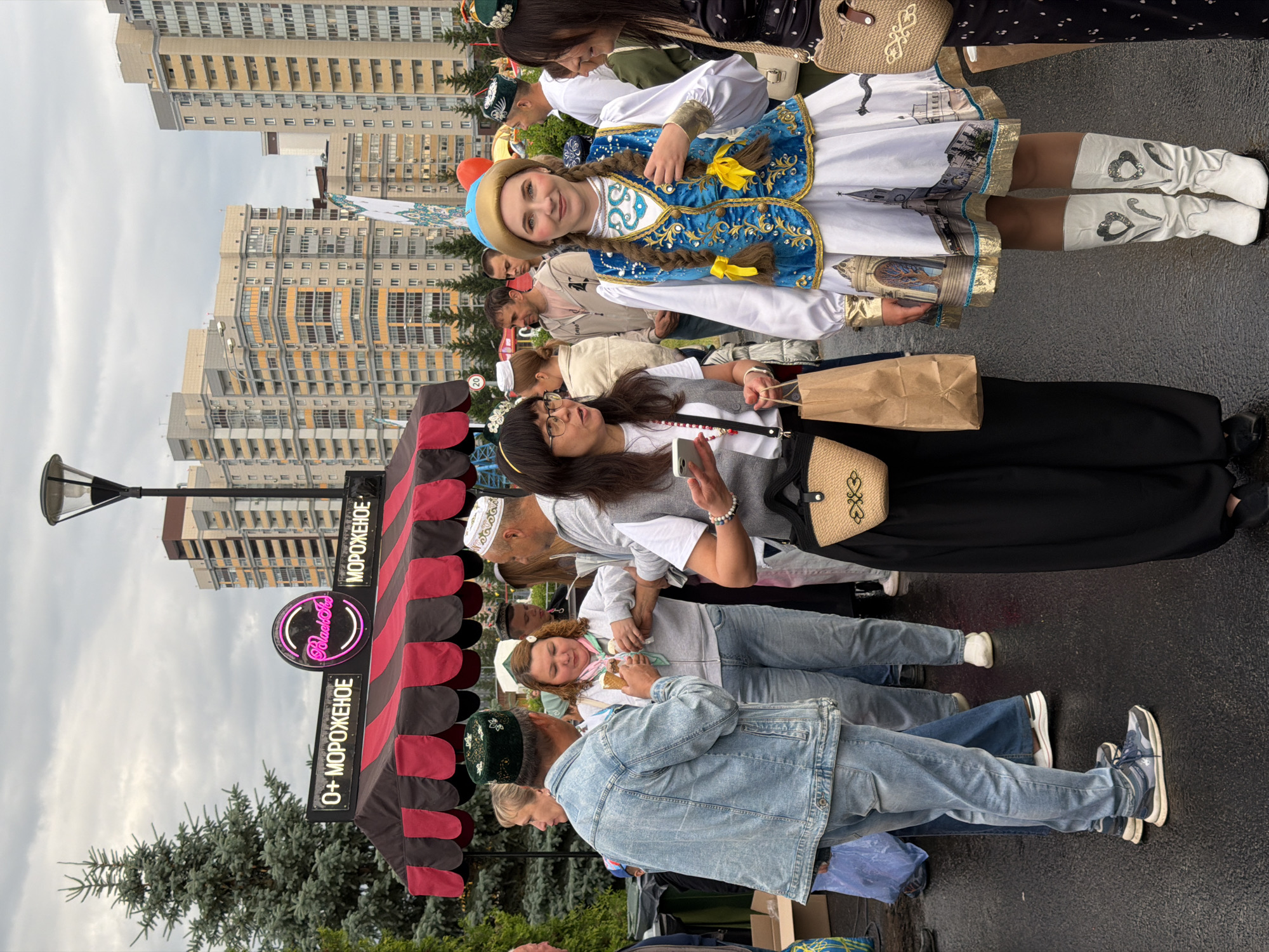In the city of Unknown, Unknown, a group of people are gathered near an ice cream stand. The stand has a striped red and black awning with neon signs that read "O+ МОРОЖЕНОЕ" and "Black Sea". Several individuals are visible, some wearing traditional headwear. A woman in a blue and gold traditional outfit, complete with braids and a gold-colored hat, stands prominently in the foreground, looking directly at the camera. She is flanked by other individuals, one of whom is holding an ice cream cone. The background features modern high-rise buildings and trees. The ground appears wet, suggesting recent rain.