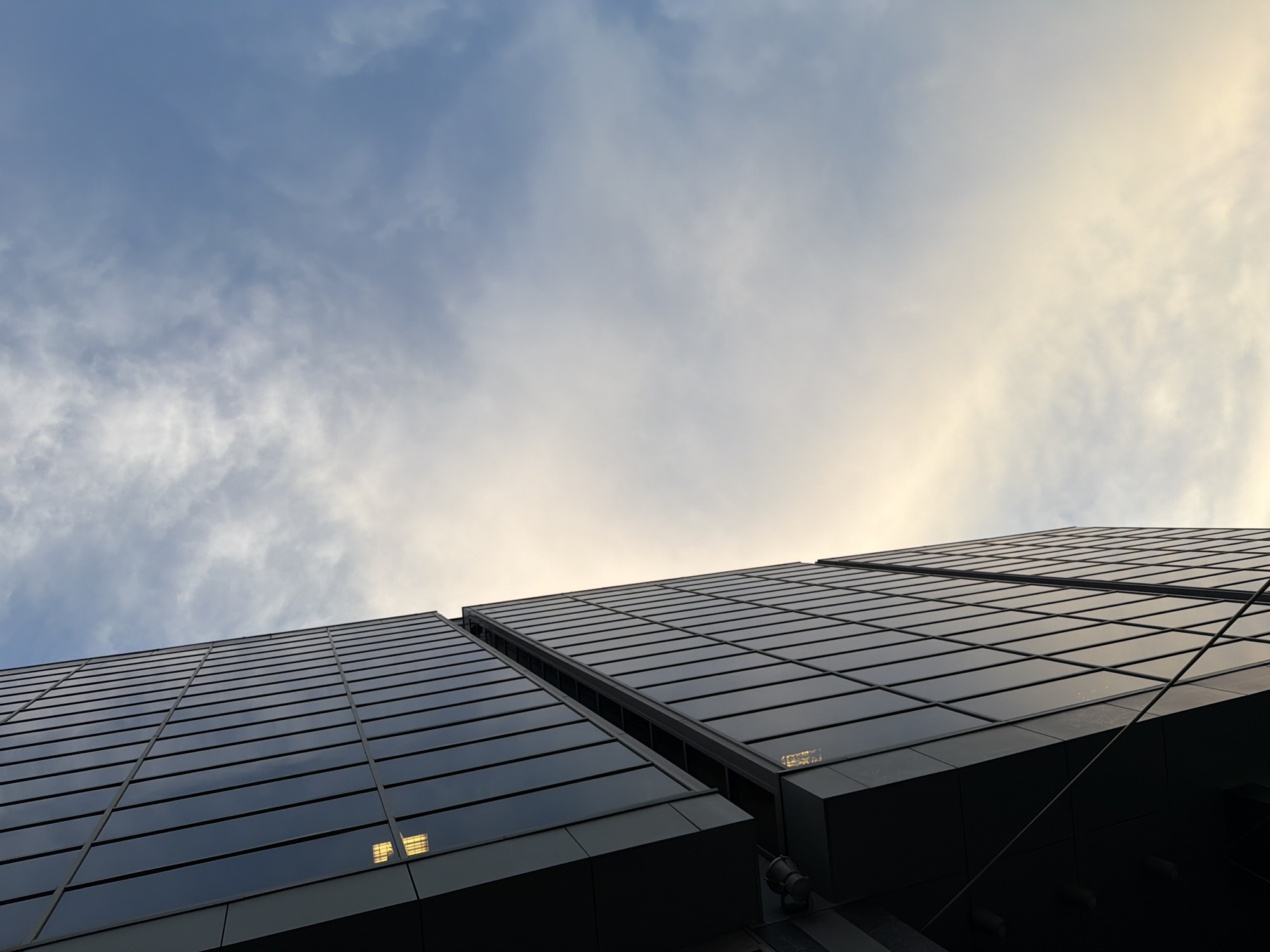 The photograph presents a low-angle view of a modern building with a predominantly glass facade, situated in an urban environment. The building's exterior is characterized by a grid-like pattern of dark, reflective windows. The sky above transitions from a pale blue to a soft, muted yellow, suggesting either sunrise or sunset. Wispy clouds are scattered throughout the sky, adding texture and depth to the scene. The angle of the shot emphasizes the height and scale of the building.