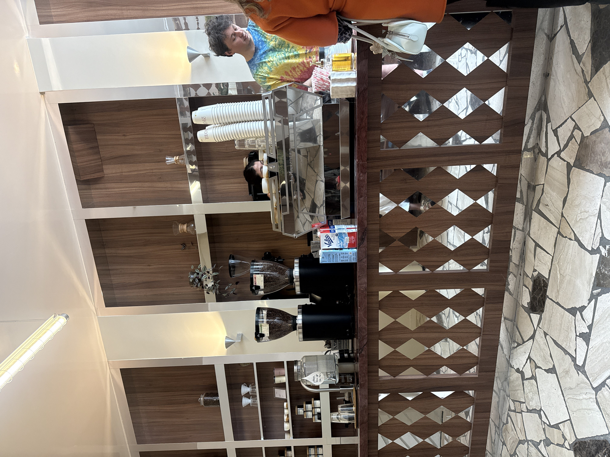 The image shows the interior of a cafe or coffee shop. A man in a tie-dye shirt and orange jacket is standing behind a counter, likely a barista serving a customer. The counter has a mirrored diamond pattern on the front and is stocked with paper cups, an espresso machine, coffee grinders, and other coffee-making equipment. Shelves behind the counter display coffee beans, various ingredients, and decorative items. The floor is tiled with irregular stone shapes. The location is possibly a cafe in Paris, France, as the setting aligns with typical Parisian coffee shop aesthetics.