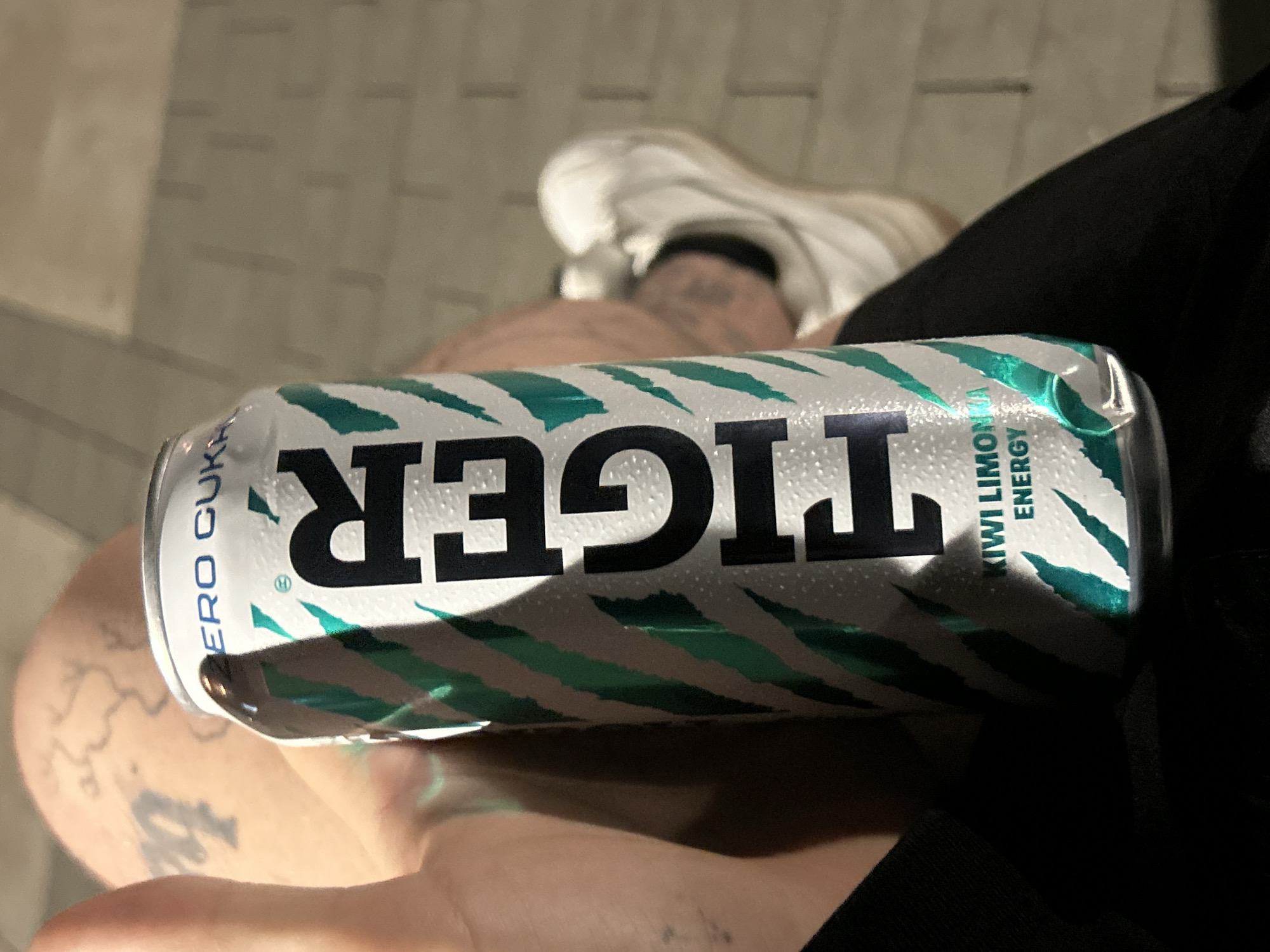 A young adult, likely male given the tattoos and clothing, is holding a can of "Tiger" energy drink. The can is silver with a green and white striped design, and the word "TIGER" is prominently displayed in black lettering. Text on the can indicates "ZERO CUKRU" and "KIWI LIMONKA ENERGY". The person is wearing black clothing and white sneakers. Tattoos are visible on the person's left arm and thigh, one appearing to be a map outline. The background consists of paved ground, possibly a sidewalk or plaza, with a brick-like texture. The image appears to have been taken outdoors in the evening or at night, given the lighting. The person's left thumb is visible in the foreground, positioned as if to support the can. The overall setting is urban, and based on the presence of the "ZERO CUKRU" text, which is Polish for "zero sugar", the location is likely Poland, specifically the city of Siedlce.