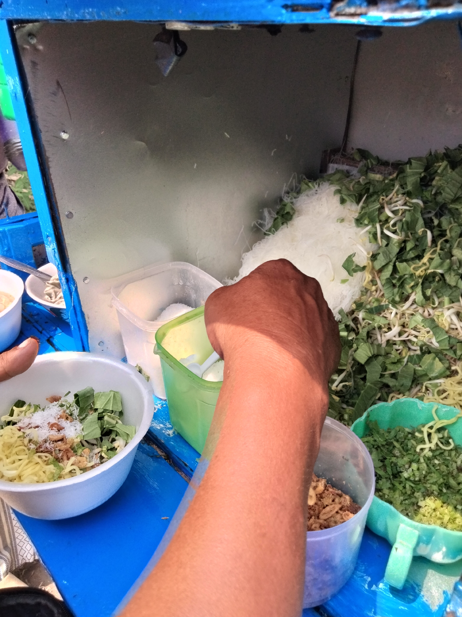 In Cariu, Indonesia, a hand is visible in the foreground of the image, reaching into a green container. The hand appears to be holding a white scoop and is about to scoop something from the container. To the left of the hand, there is a white bowl filled with noodles, chopped green vegetables, and white flakes, likely salt or sugar. Adjacent to the green container is a clear plastic container with a white granular substance. Behind these containers, a pile of white noodles and chopped green vegetables are visible, along with some bean sprouts. The scene takes place outdoors, with a bright, sunny environment suggested by the lighting and shadows. The blue painted wooden surface indicates a market stall or food vendor setup.