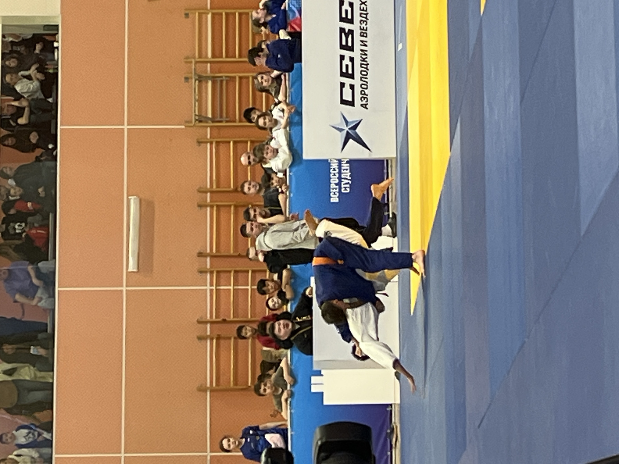 A judo match is taking place. A judoka wearing a white uniform and a black belt is lifted by a judoka wearing a dark blue uniform with an orange belt. The judoka in the dark blue uniform is in the process of throwing the judoka in white. There are spectators in the background. The background shows a beige wall with gymnastic ladders. A banner is visible in the background with Russian text and a star. The scene is presumably in Krasnoyarsk, Russia.
