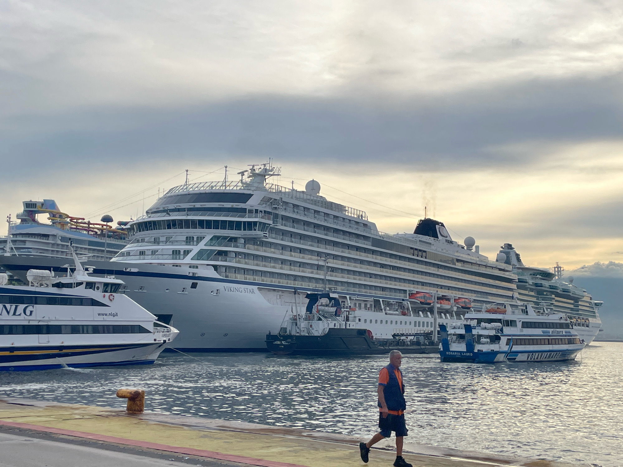 In the city of Naples, Italy, a man in an orange vest and dark shorts walks on a pier. Behind him, several large cruise ships are docked in the harbor. The closest ship, a white ferry with blue and yellow stripes, has "NLG" and "www.nlg.it" printed on its side. Further back, a massive white cruise ship named "VIKING STAR" dominates the scene, with several decks of cabins visible. Another smaller white vessel with blue trim and the name "VOLAVIAMARE" is docked next to the Viking Star. The sky is overcast, with soft light suggesting either early morning or late afternoon. The water in the harbor is choppy with reflections of the ships and sky. A rusted mooring bollard is prominent on the pier in the foreground.