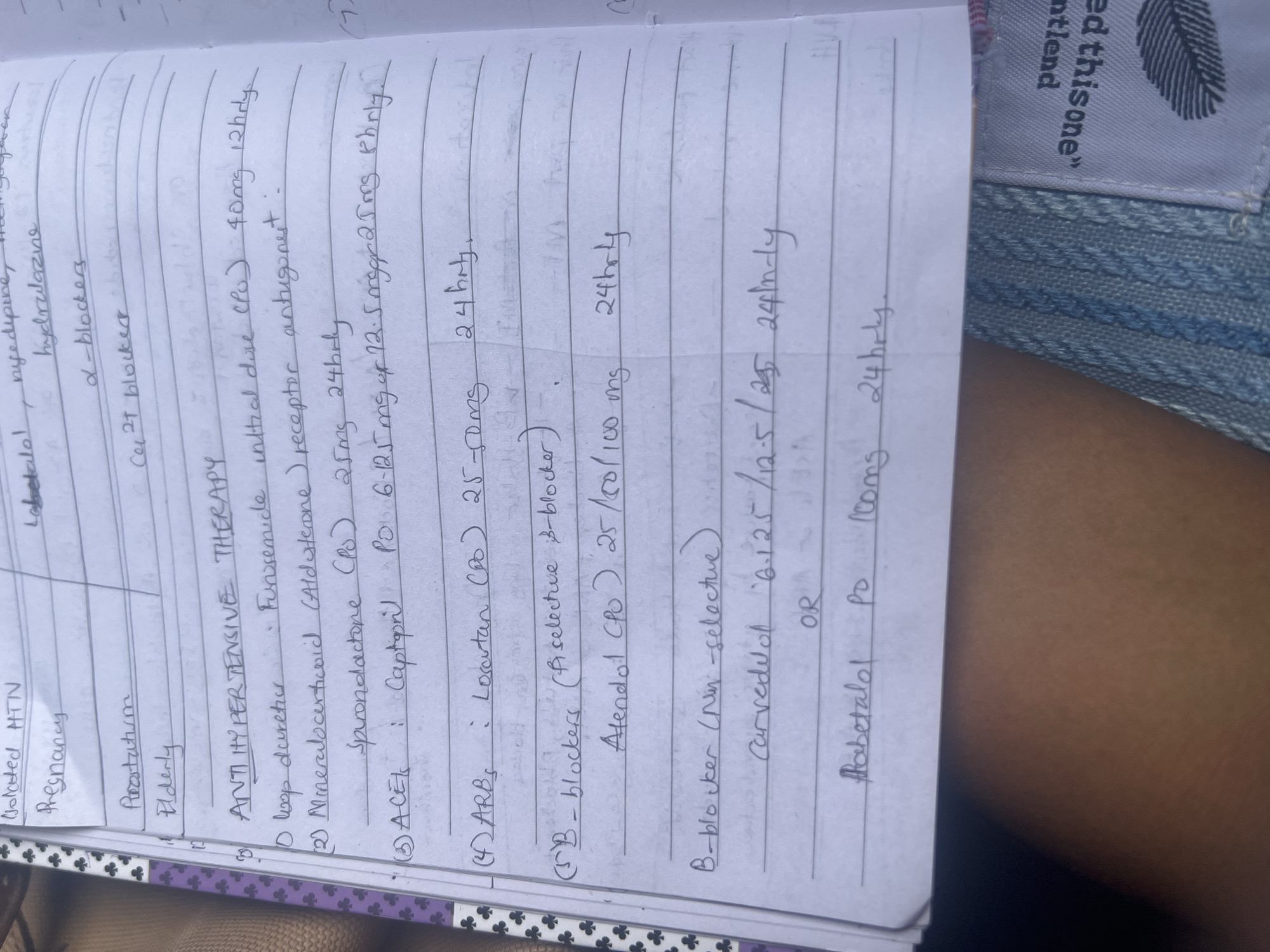 The image shows a notebook page with handwritten notes on antihypertensive therapy. The notes cover various drug classes, including loop diuretics (furosemide), mineralocorticoid receptor antagonists (spironolactone), ACE inhibitors (captopril), ARBs (losartan), and beta-blockers (atenolol, carvedilol, labetalol). Dosages and frequencies of administration are also listed. The notebook is resting on what appears to be a lap covered by blue denim clothing, suggesting an outdoor setting. A tag with text "ed this one" is partially visible on the top right corner. There is no information to determine if this image was taken in Dar es-Salaam, Tanzania.