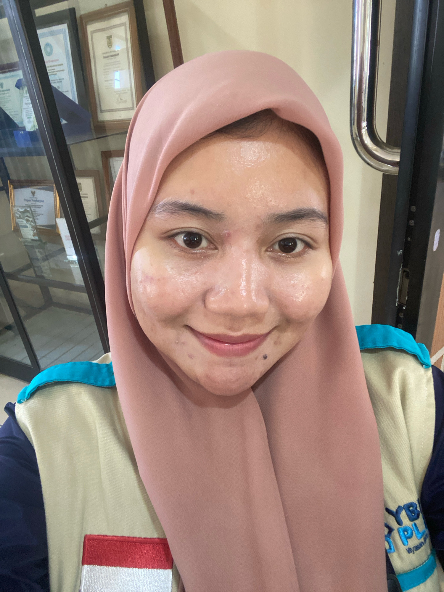 A young woman wearing a pink hijab and a tan vest with blue trim is posing for a selfie in front of a display case filled with framed documents. She has acne on her face and is smiling. The background includes a dark doorway with a silver handle and a framed document with Indonesian text. The woman is wearing a vest with an embroidered logo that includes the letters "YB" and "PL" in blue. There is also a patch of red and white fabric on the vest, which resembles the Indonesian flag. The scene appears to be indoors, possibly in an office or educational institution in Banjarbaru, Indonesia, given the presence of framed certificates and the Indonesian flag motif.