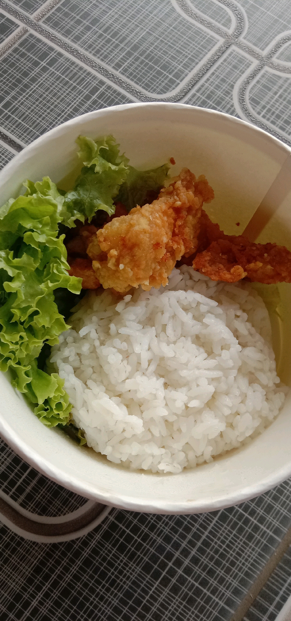 A white paper bowl filled with food rests on a patterned tablecloth. The bowl contains a generous portion of white rice, topped with several pieces of golden-brown fried chicken. To the left of the rice, a cluster of fresh green lettuce leaves adds a touch of color. The background is a dark gray and white plaid pattern of the tablecloth, with a subtle beige stripe bordering the table's edge. The lighting suggests an indoor setting, likely a meal served on a table. The specific location in Cangkring Rembang, Indonesia, is not visible in the image, as the scene focuses solely on the food.