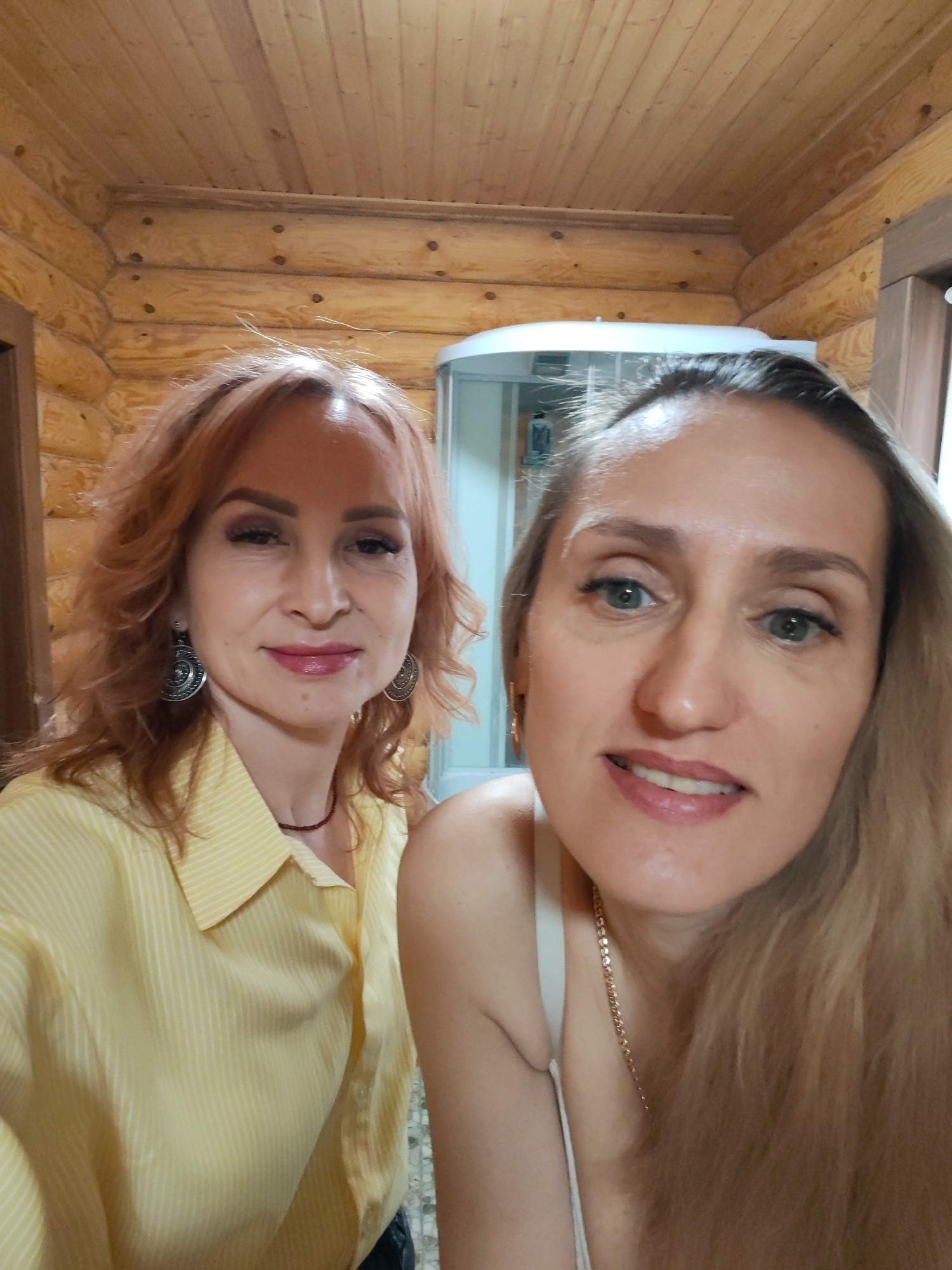 Two women pose indoors, possibly in a bathroom with log cabin-style walls. The woman on the left has curly red hair, silver earrings, and wears a striped yellow button-down shirt with a beaded necklace. The woman on the right has long blonde hair and wears a white top with a gold necklace. In the background, a shower stall is visible. The setting appears to be in a building made of logs. Based on the log cabin style and visible shower stall, it is likely a house or a cottage. The location provided is Старое Михайловское, Russia.