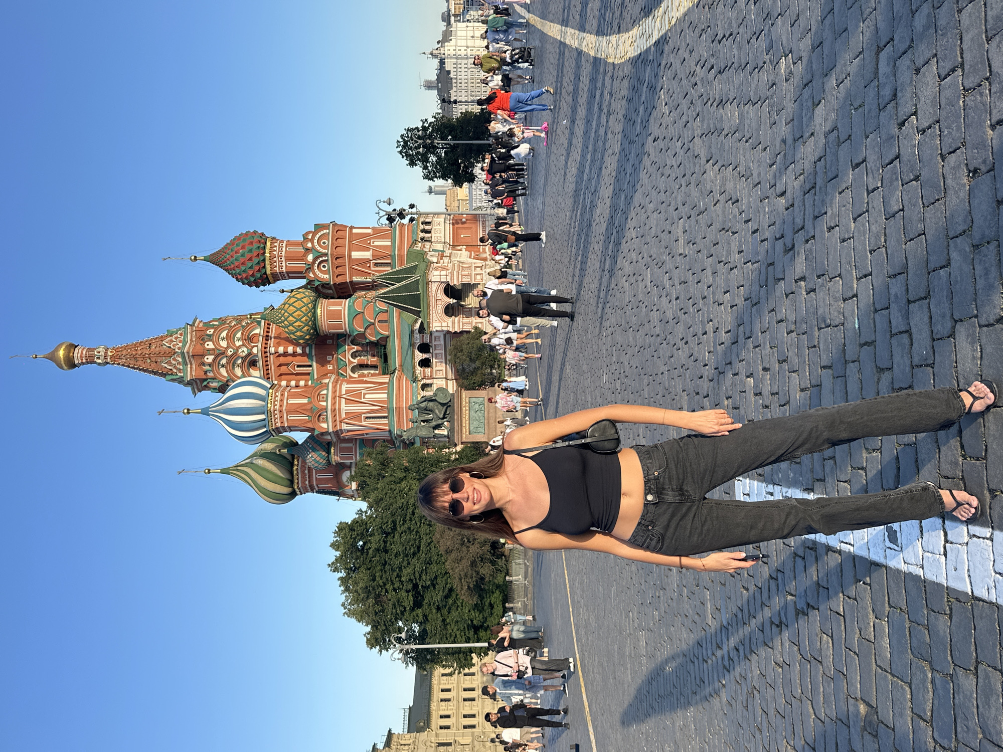 A woman with long brown hair and sunglasses poses for a photo in front of Saint Basil's Cathedral in Moscow, Russia. She is wearing a black tank top, dark wash jeans, and black flip-flops. The cathedral, with its iconic onion domes, is visible in the background, bathed in sunlight. The ground is paved with cobblestones, and other tourists are seen walking around the area. The sky is clear and blue.