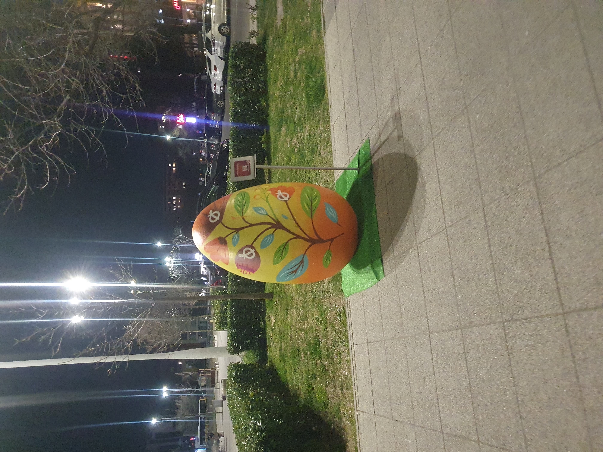 The image captures a large, decorated Easter egg sculpture placed on a small green mat, situated on a tiled sidewalk in Plovdiv, Bulgaria. The egg features an orange background with painted designs including branches, flowers, and owls. A metal stand with a small square sign supports the egg. The backdrop includes a grassy area with trimmed hedges, bare trees, and streetlights illuminating the scene. Further in the background, blurred lights indicate the presence of buildings and possibly cars. The image was likely taken at night.