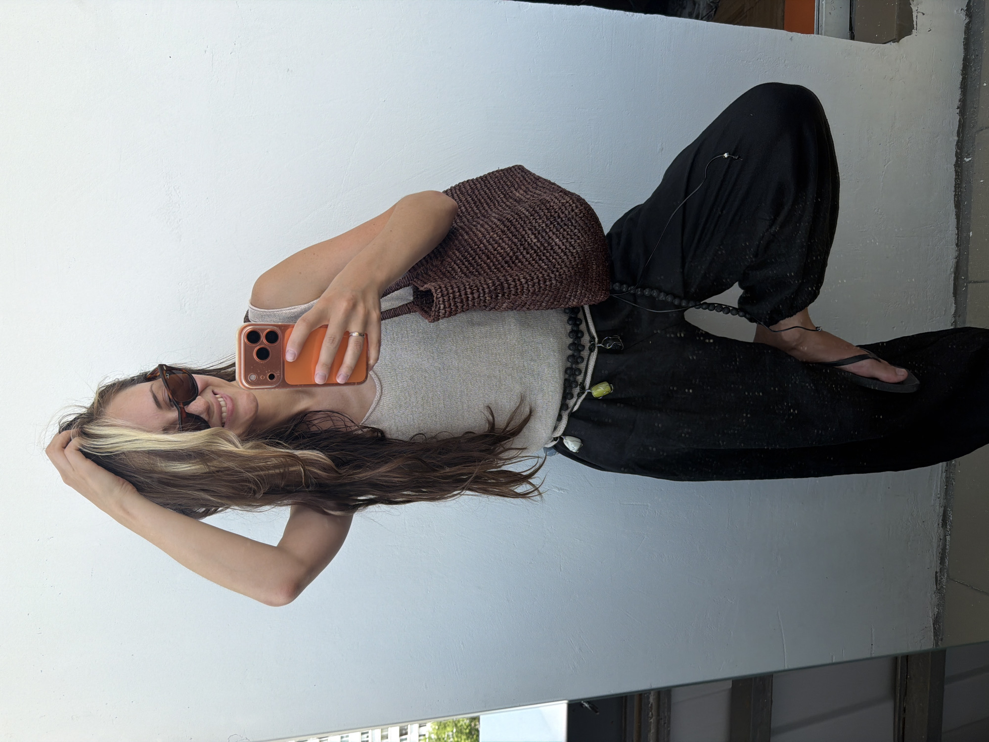 A woman stands in front of a white wall, taking a mirror selfie. She is wearing a sleeveless beige top, black baggy pants, and flip-flops. She has long, dark hair with blonde streaks and is wearing sunglasses. A brown woven bag is slung over her shoulder. She is holding an orange smartphone in her right hand and her left hand is in her hair. A beaded necklace is draped around her waist and another hangs down from her neck. The background appears to be outdoors, with a hint of greenery and buildings visible in the reflection of a mirror on the left side of the image. The lighting suggests it is daytime. The environment is a simple, outdoor urban setting, likely a balcony or patio in Moscow, Russia.