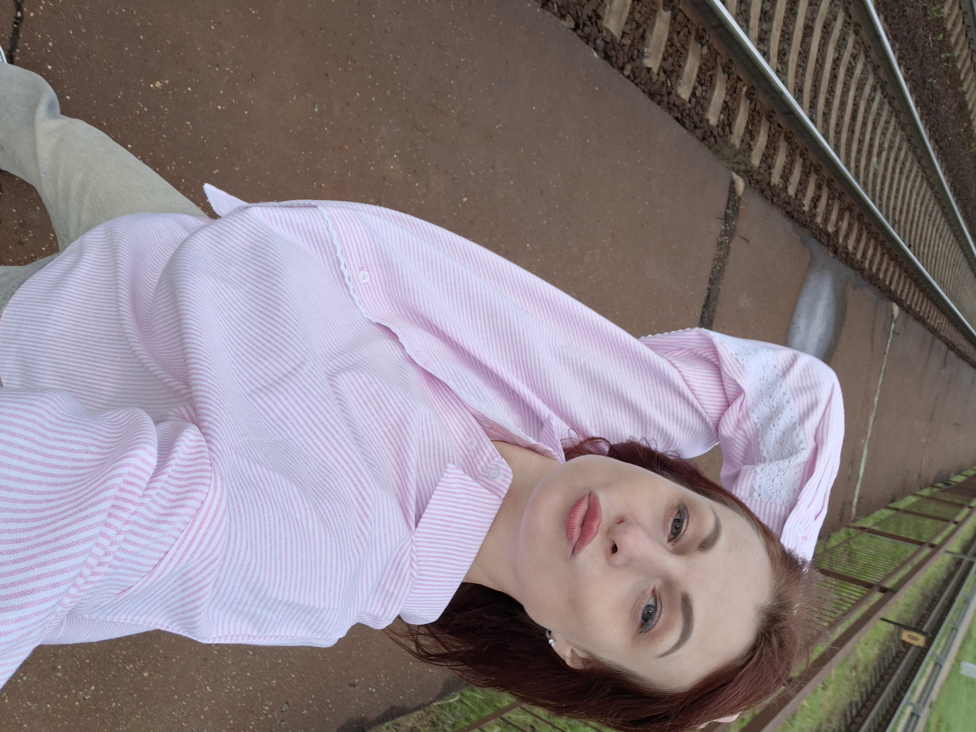 A woman with reddish-brown hair and light-colored eyes, wearing a pink and white striped button-down shirt and light-colored pants, lies on her back on a brown, gritty surface next to railroad tracks. Her head is tilted to the right, and her right arm is bent with her hand behind her head. Her lips are painted a rosy pink. The background consists of railroad tracks, with green grass visible beyond the tracks, suggesting an outdoor setting. The lighting appears to be natural, indicating it might be daytime. The overall scene conveys a relaxed or contemplative mood. No visible text is present in the image.