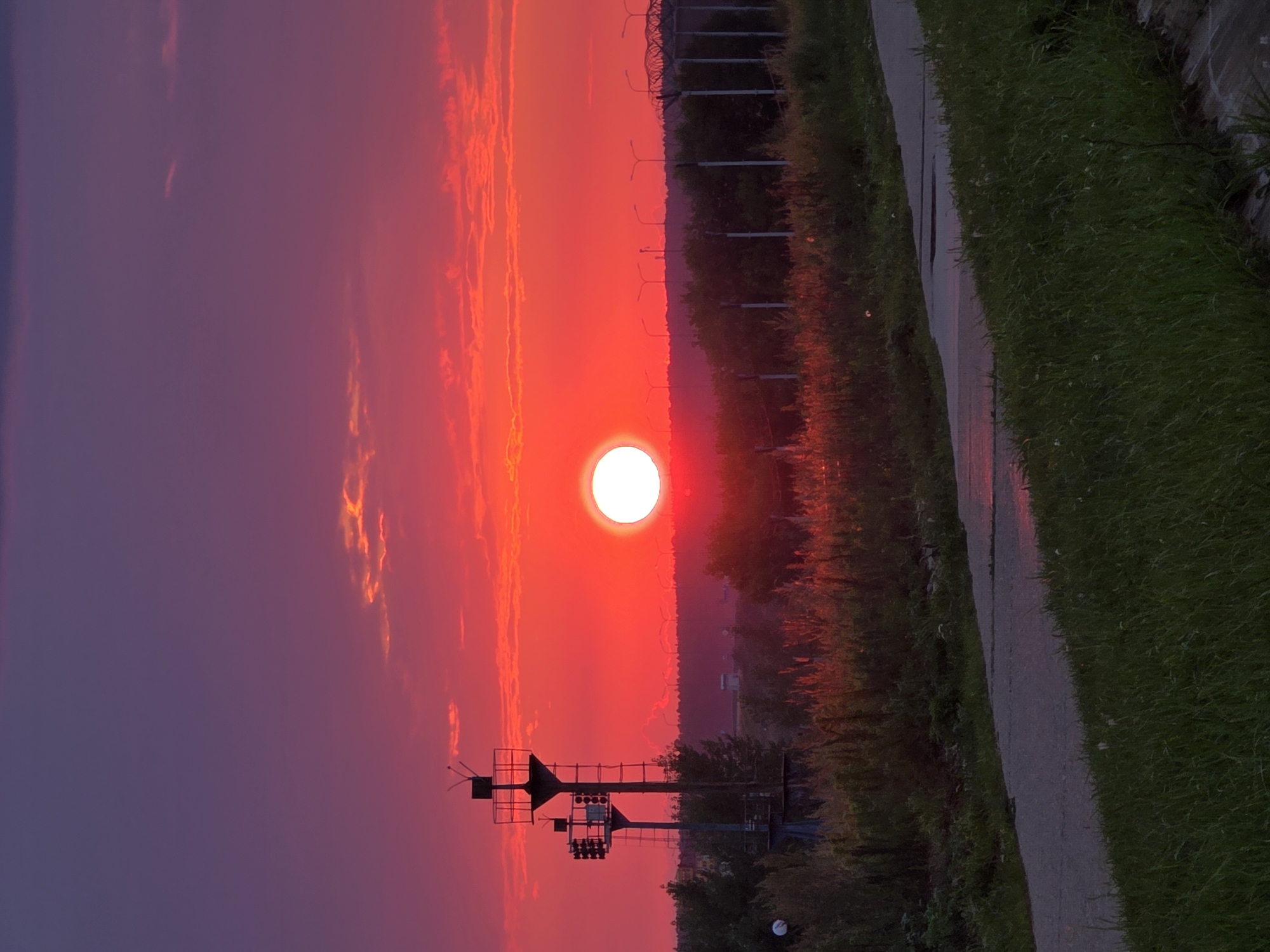 The image captures a vibrant sunset over the city of Chaikovsky, Russia. The sky is a gradient of deep purple at the top, transitioning to intense shades of red and orange near the horizon. A brilliant, bright orange sun is prominently positioned in the center of the frame, its light casting a warm glow across the scene.
In the foreground, a paved path, possibly a walkway or road, curves through lush green grass. The path appears slightly wet in places, reflecting the sunset's colors. To the left of the path, two tall industrial-style structures with platforms and lights stand silhouetted against the fiery sky. These structures are likely navigational aids or part of an industrial facility.
Beyond the path and structures, a dense line of trees forms the horizon, with some taller structures and utility poles visible interspersed. The overall impression is one of a dramatic natural phenomenon occurring in an urban or semi-industrial environment. No people are visible in the image.