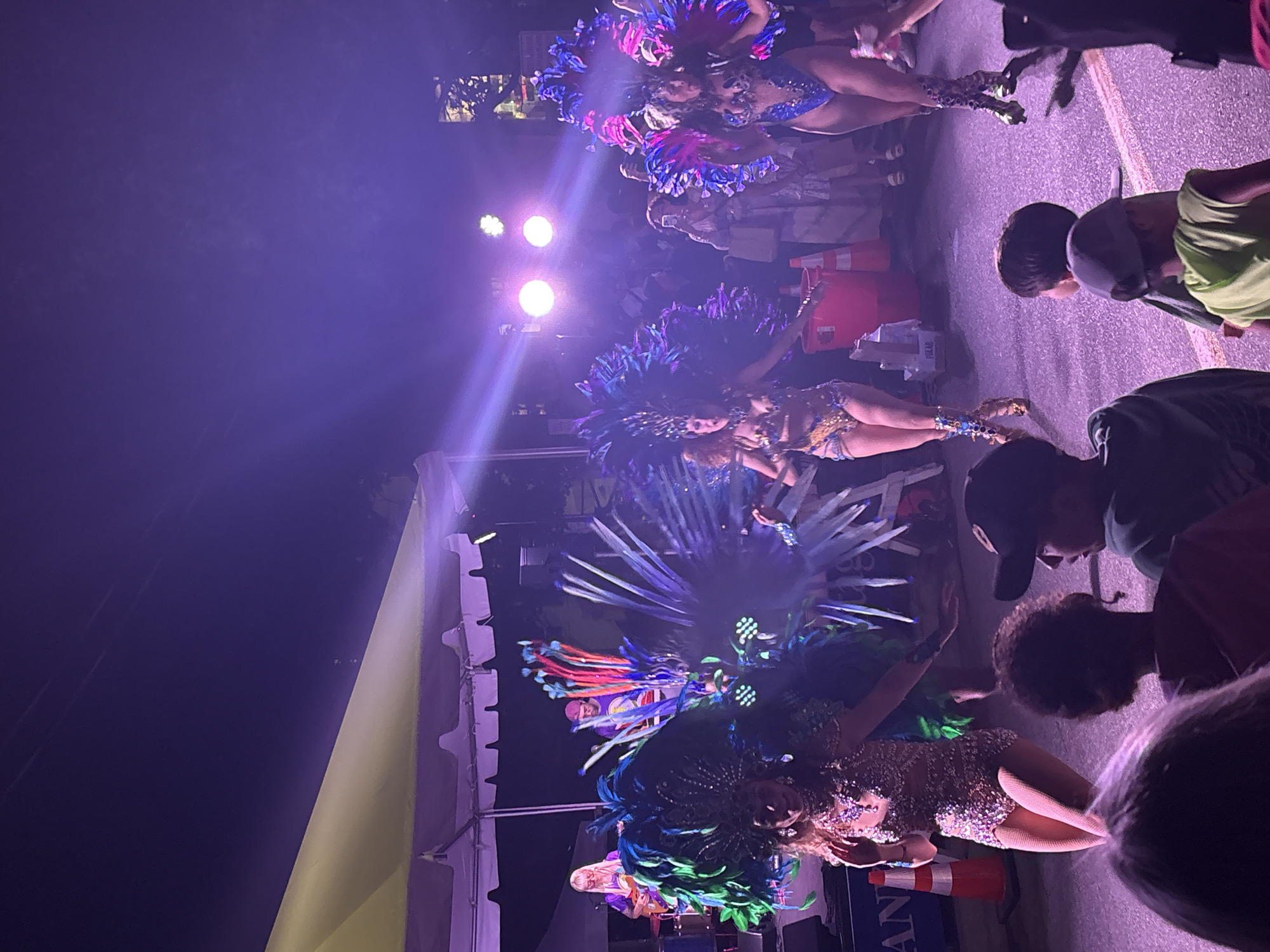 In Provincetown, Massachusetts, a nighttime outdoor event features performers in vibrant, feathered costumes on a street stage. The stage is set under a large, light-colored tent, illuminated by purple and pink stage lights, creating a festive atmosphere. Two main performers, adorned with elaborate headdresses and sequined outfits, are the focus. The performer on the left wears a shimmering costume with green and blue feathers, while the performer on the right is in a blue sequined swimsuit with pink and blue feathers. Both have on high-heeled boots. In the background, a band is visible with a guitarist and a keyboard player. Spectators, including children, are gathered at the front of the stage, watching the performance. Some individuals in the crowd are holding up phones to capture the event. Traffic cones and a red bucket are visible near the stage, suggesting a temporary setup for the event. The overall scene depicts a lively celebration with a strong visual emphasis on the performers' dazzling attire and the colorful lighting.