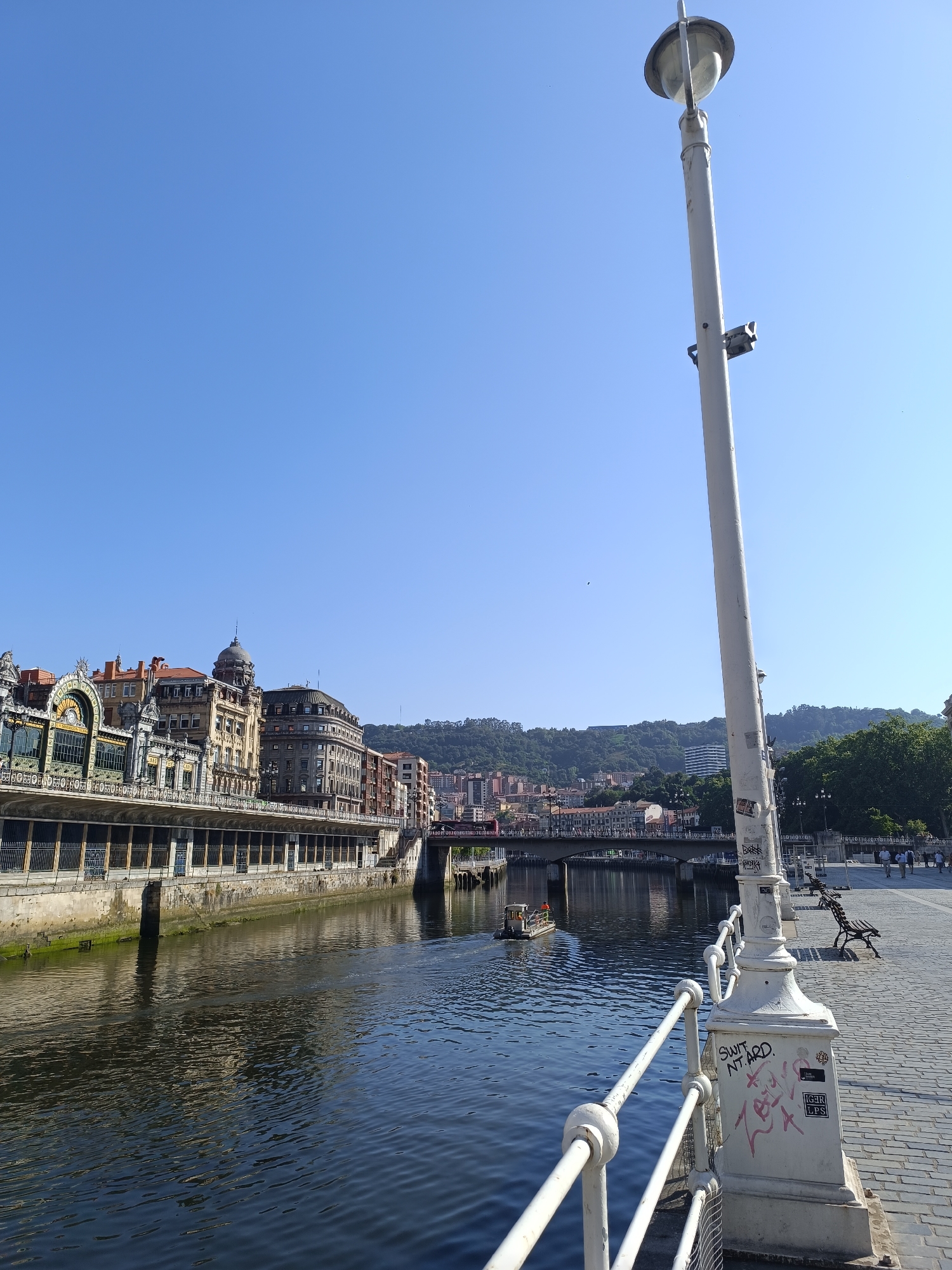 The image shows a view of the Nervión River in Bilbao, Spain, on a clear day. In the foreground, a white lamppost with graffiti stands to the right. A white railing runs along the waterfront. To the left, the river flows with reflections of the buildings on the opposite bank. A small boat is visible on the water, heading towards a bridge in the distance. The opposite bank features a row of historic buildings with ornate facades, characteristic of Bilbao's architecture, and a hill covered in trees and buildings rises behind them. A paved walkway with benches is visible along the waterfront in the middle ground, where several people are walking. The sky is a clear, bright blue.