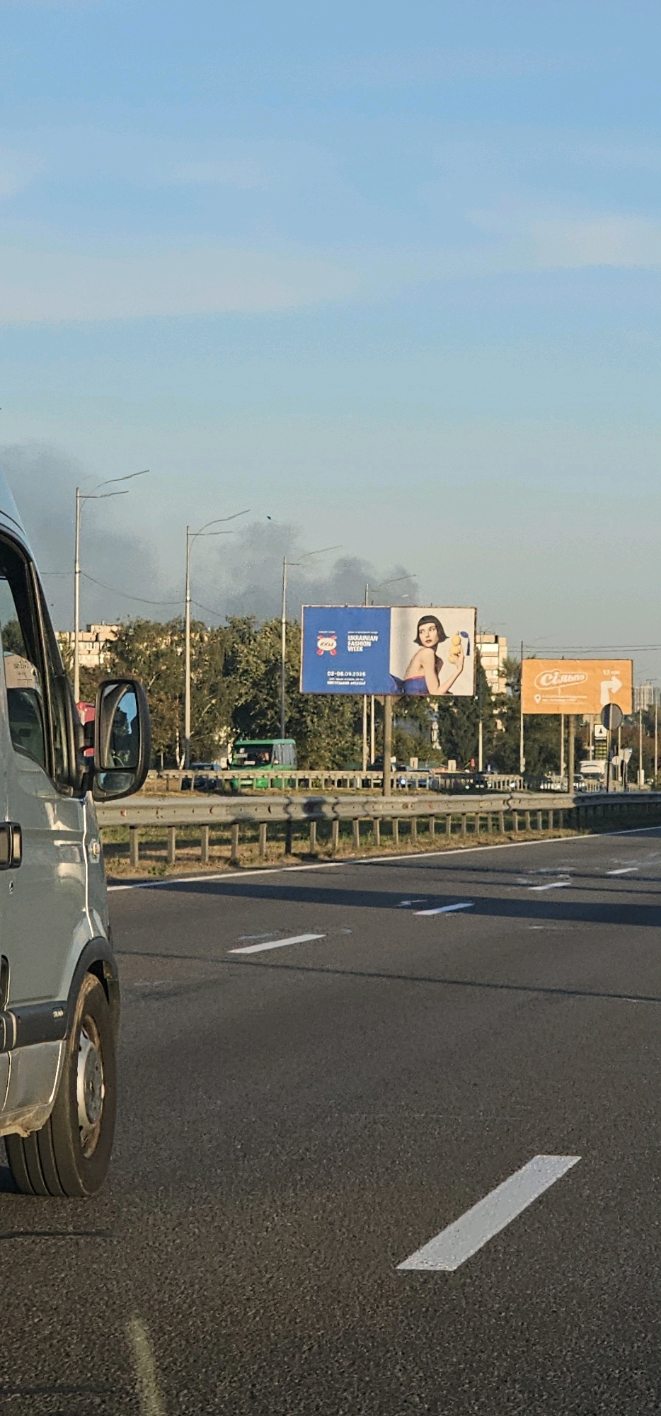In this image, taken from the perspective of a vehicle on a highway in Kyiv, Ukraine, several elements are visible. The sky is a clear blue with some light clouds. Smoke plumes are rising in the distance, suggesting a fire or industrial activity. Two billboards are prominent in the background. The left billboard is blue and features a woman holding lemons, with text that appears to be an advertisement for "Ukrainian Fashion Week" with dates 03-06.09.2020. To the right of this is an orange billboard with text, likely another advertisement. The highway itself has clear lane markings. Trees and buildings line the side of the road. In the foreground, the side and wheel of a light-colored van are visible, indicating the viewpoint. A green bus is seen driving on the highway in the middle ground.