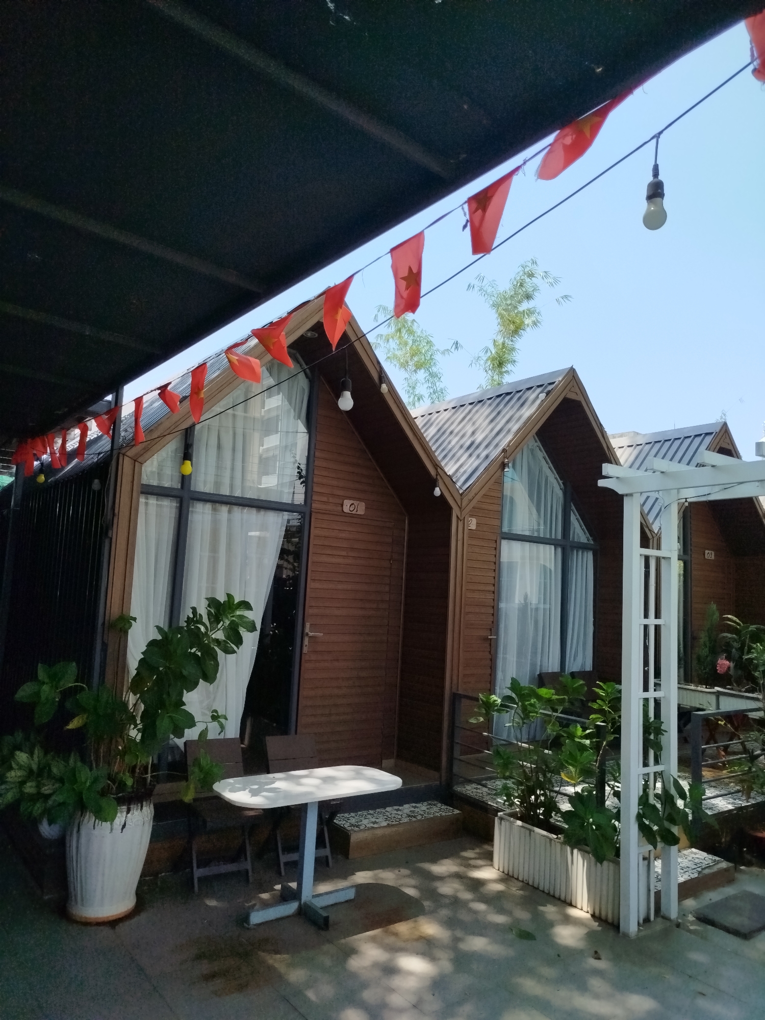 The image captures a row of small, wooden A-frame cabins or bungalows under a shaded area, likely in an outdoor cafe or resort setting. Vietnamese flags are strung above the cabins as decorative elements. The cabins have large, triangular windows covered with white curtains. Each cabin has a number. A white, metal frame structure is to the right of the cabins, possibly part of a pergola or decorative feature. There are tables, chairs, and potted plants outside the cabins, suggesting a relaxation or dining space. The scene seems to be in an urban area. The specific location within Ho Chi Minh City, Vietnam, cannot be determined from this image alone.