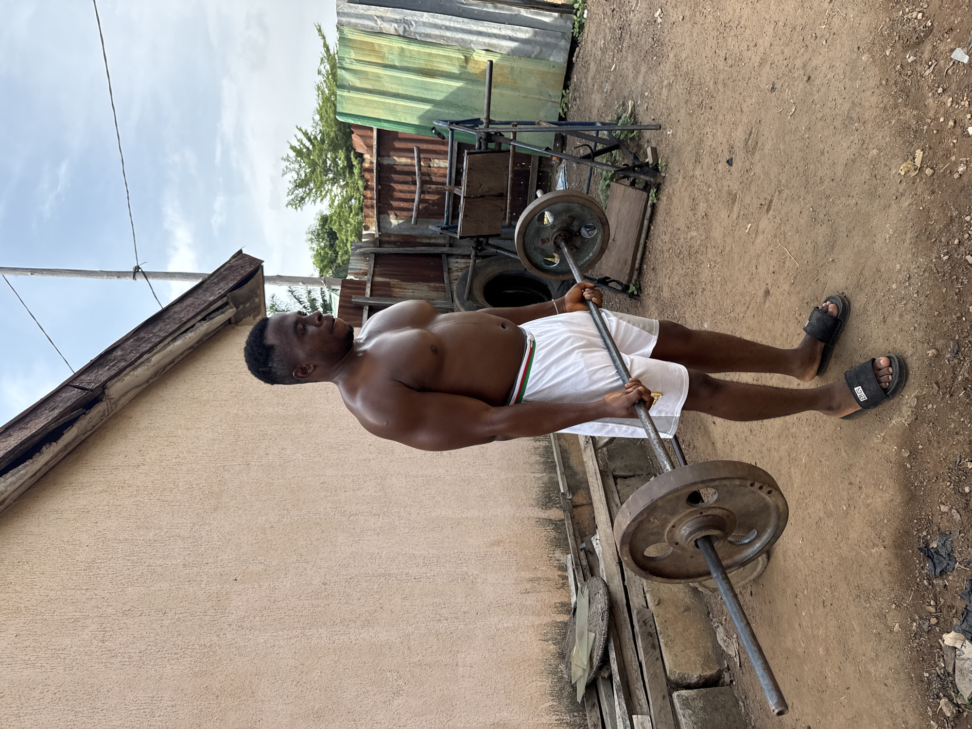 A shirtless man in white shorts and sandals is seen holding a barbell with weights in an outdoor location. The setting appears to be a compound or backyard in Akwanga, Nigeria, as suggested by the corrugated metal fencing, weathered building exterior, and dirt ground. The man is positioned in the center of the frame, engaging in what seems to be a weightlifting exercise. Behind him, there's makeshift workout equipment and other miscellaneous items scattered around the area. The overall environment suggests a basic and utilitarian space.
