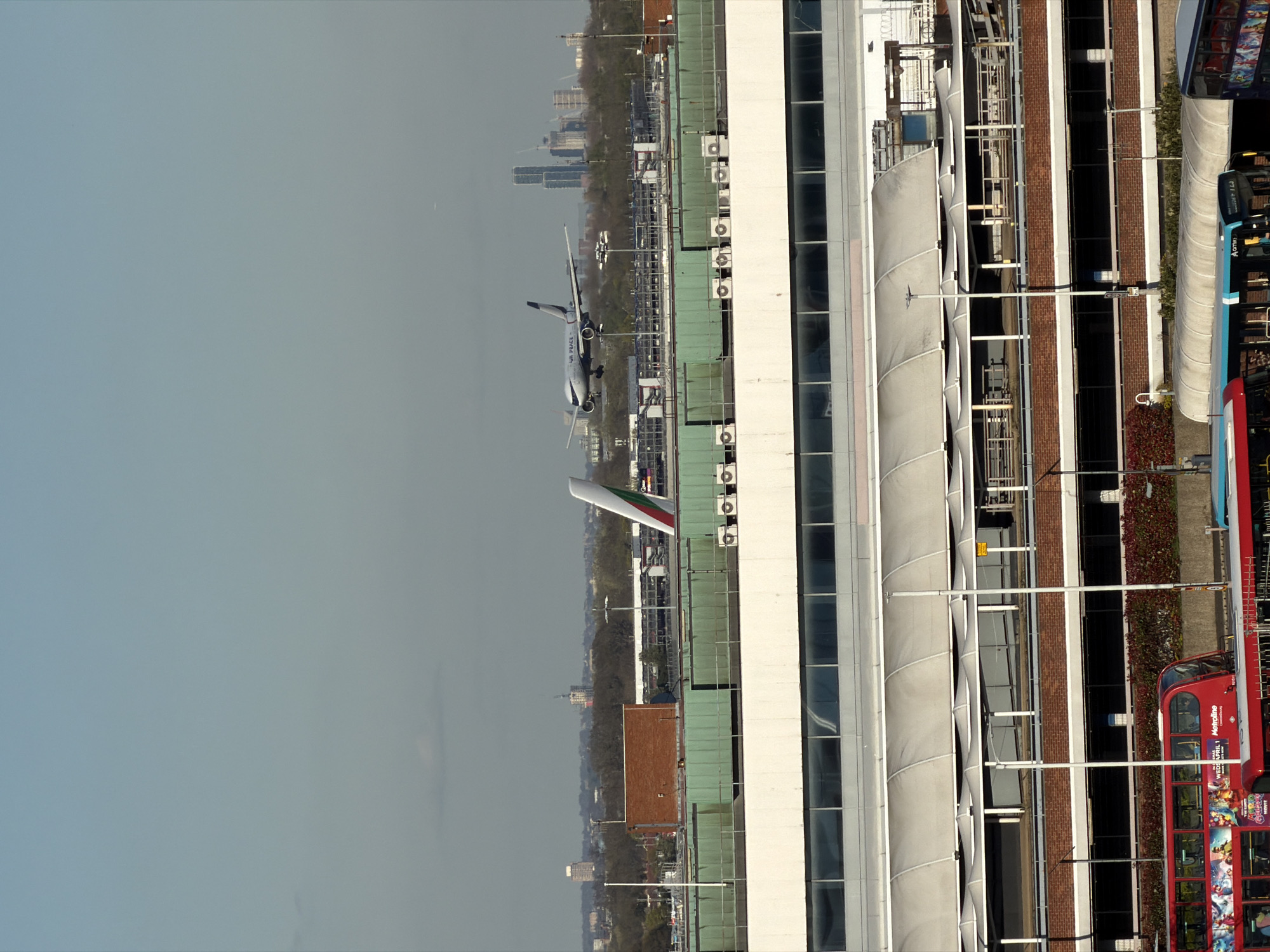 The image captures a low-angle shot in London, United Kingdom, featuring an airplane descending towards Heathrow Airport. The plane, painted white with a green, white, and red tail fin, is flying over rooftops and trees. In the foreground, part of a tall building with white and grey vertical lines is visible on the right side of the frame, along with sections of brick and glass. Adjacent to the building, a red double-decker bus is parked on the road. In the background, the city skyline is visible through a hazy atmosphere. The sky is a pale greyish-blue.