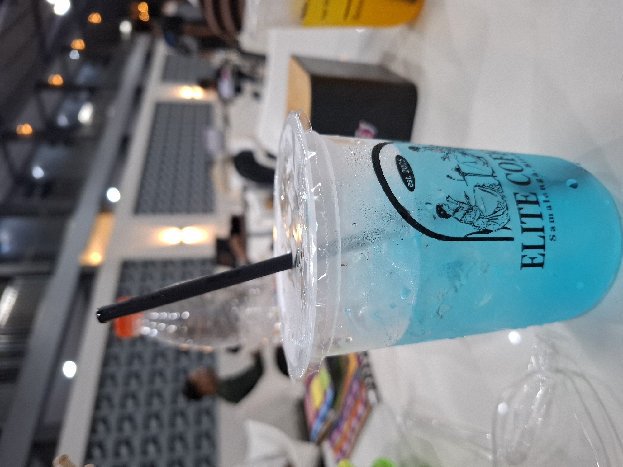 A close-up, eye-level, indoor shot shows a clear plastic cup filled with a bright blue beverage and ice, placed on a white table. A black straw protrudes from the cup. The cup has a white label with black text and two figures depicted in silhouette, one seated at a table and another standing. The text on the label reads "est. 2025" and "ELITE COF" with "Samalanga. Bir" below. The background is blurred, showing a dimly lit interior of what appears to be a cafe or restaurant. To the left of the cup, a stack of colorful magazines or books is visible. In the far background, out of focus, are people and a decorative wall with a patterned texture. The overall ambiance suggests a casual setting.