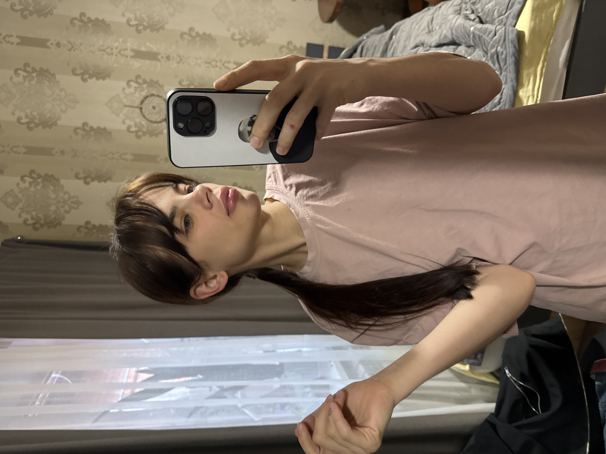 A woman is taking a selfie with her iPhone in an indoor setting. The phone case is white with a black border, and she is holding the phone with her left hand. She has brown hair and is wearing a pink t-shirt. The background includes a wall with a floral pattern, a window with curtains, and a bed with a gray comforter. There is no verifiable information to conclude that this image was taken in the city of Rostov-on-Don, Russia.