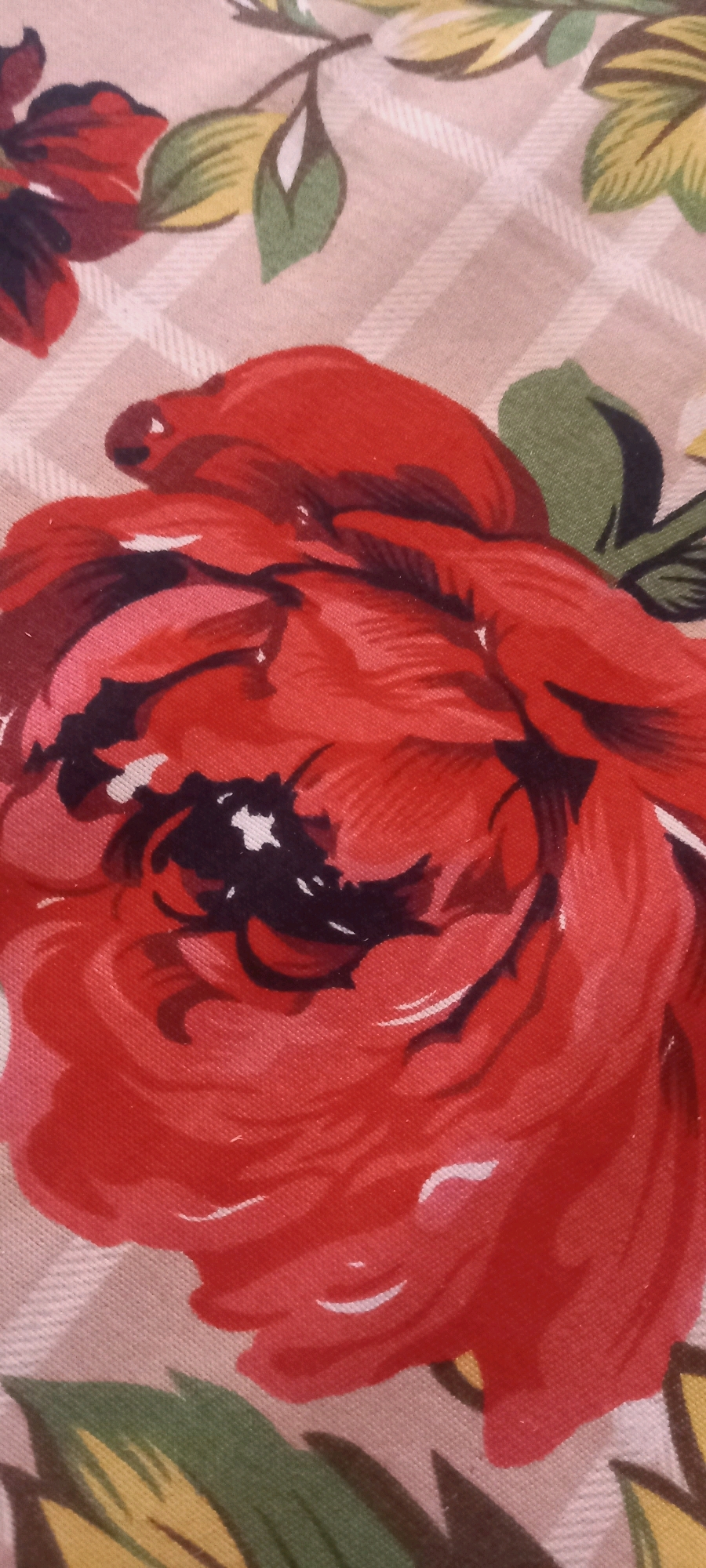 The image displays a close-up view of a fabric with a floral pattern. The dominant element is a large, red flower, likely a rose, with detailed petals painted in various shades of red, pink, and dark maroon. Some black and white accents are visible within the flower's center. The flower is set against a background of a light beige fabric with a subtle plaid or checkered pattern. Green leaves with yellow and brown accents are also visible, scattered around the flower. The fabric appears to be a textile material, possibly cotton or a blend, and the pattern suggests it could be from a garment, upholstery, or decorative item. The image's perspective is a top-down, slightly angled view of the fabric. The location in Pakpattan, Pakistan is not discernible from the visual content of the image.