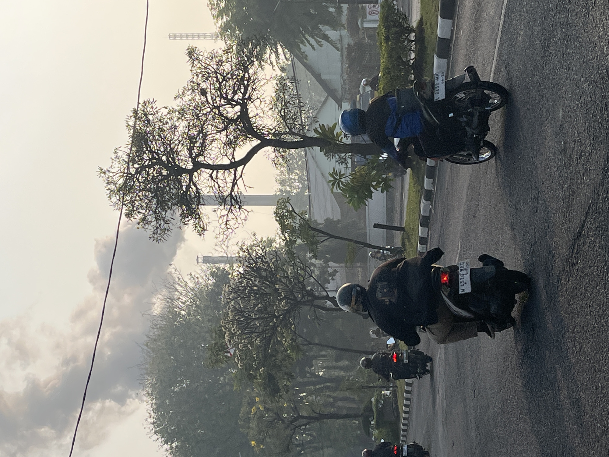 In the city of Candi, Indonesia, a hazy, possibly smog-filled sky looms over a road where several individuals are riding motorcycles. In the foreground, two individuals on motorcycles are prominently featured, viewed from behind. The rider on the left wears a dark jacket and tan pants, while the rider on the right is clad in a dark jacket and blue pants, both wearing helmets. The motorcycle on the right displays a license plate with the characters "L 2452 MK". Other motorcyclists are visible further down the road. Trees with sparse foliage line the sides of the road, and industrial structures with smoking chimneys are visible in the background, suggesting the presence of factories or industrial facilities. The overall atmosphere is muted due to the haze, with a pale, overcast sky.